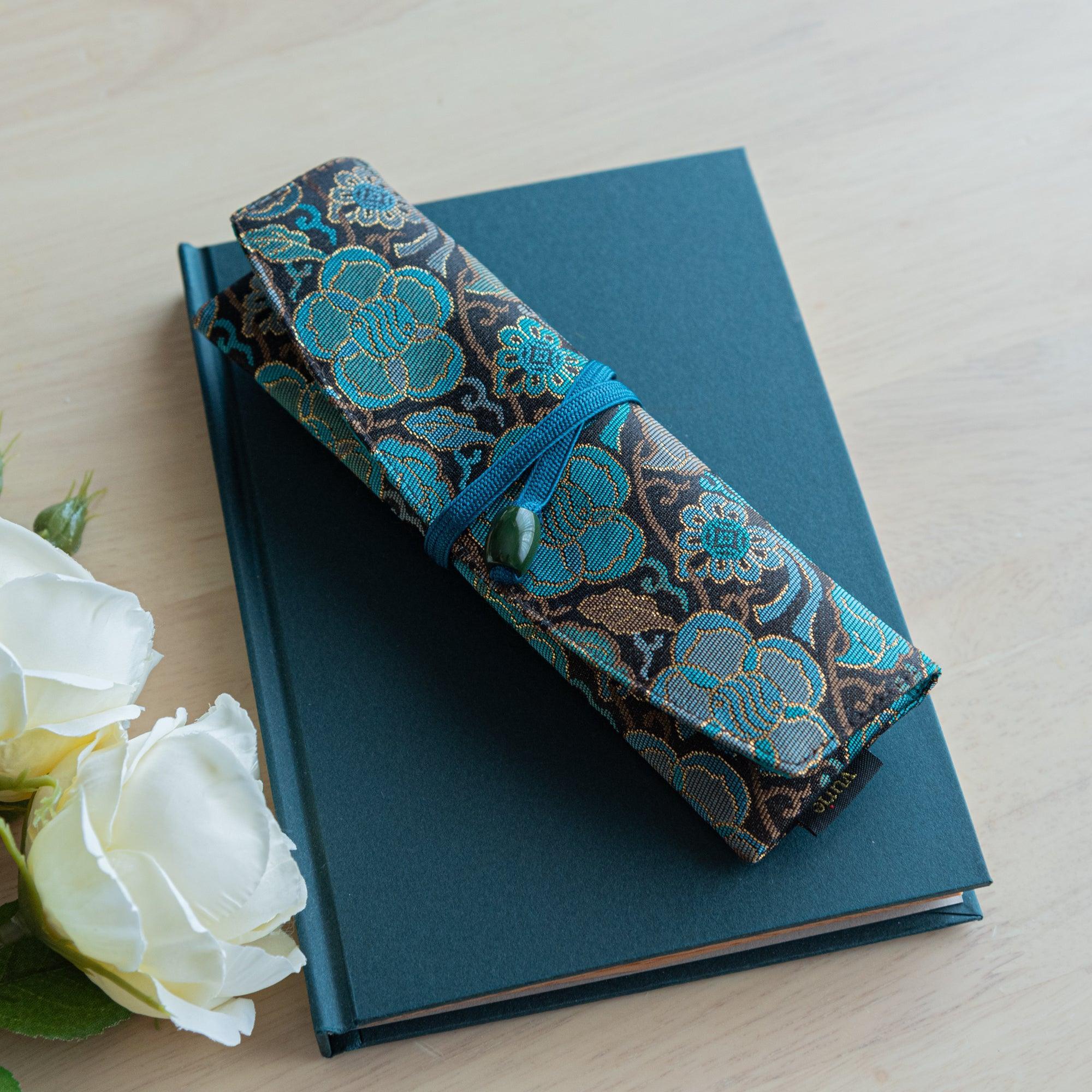 A folded teal and gold Japanese fountain pen case, resting on a dark teal notebook, with white roses nearby on a light wooden surface.