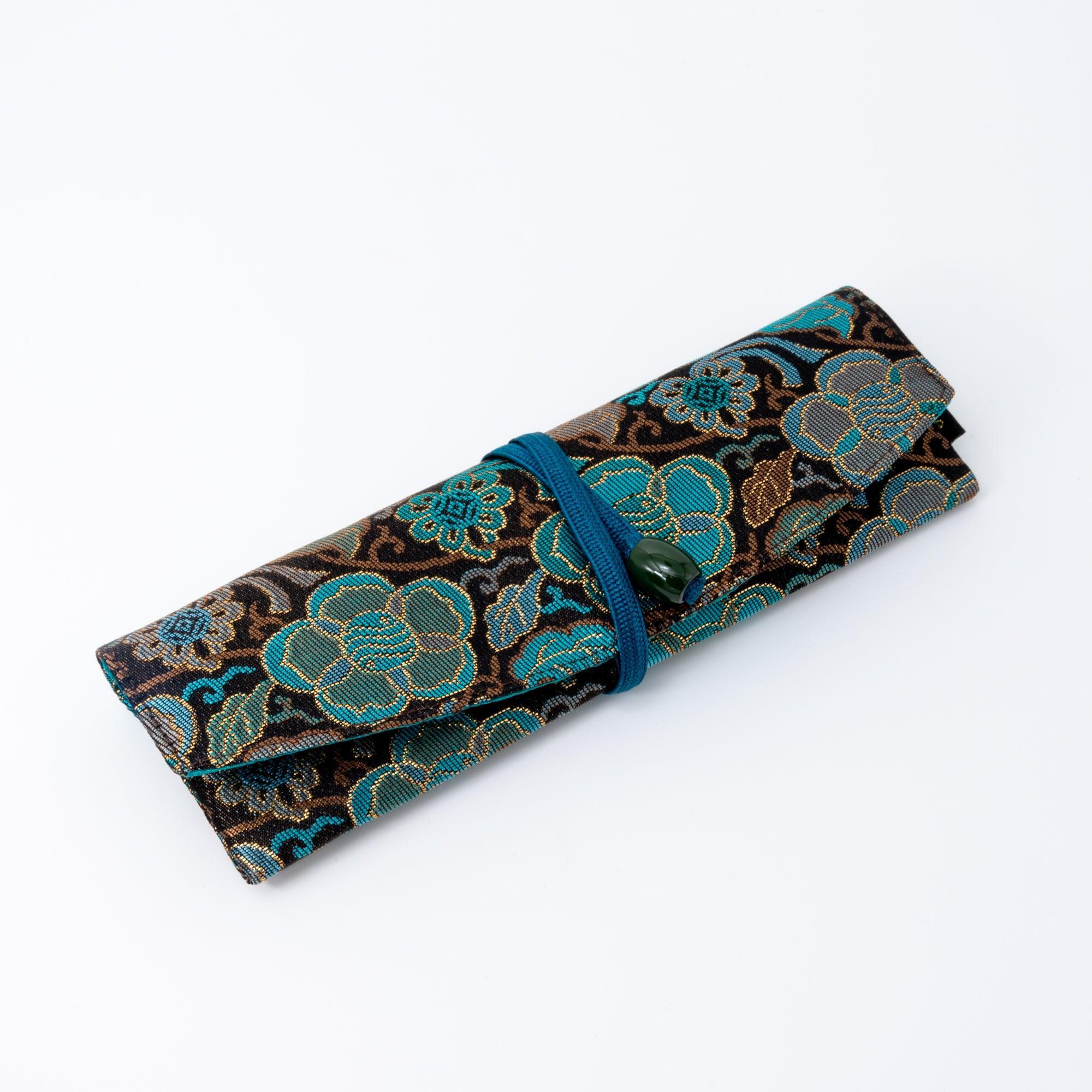 A compactly folded teal and gold Japanese fountain pen case of Nishijin silk, tied securely with a blue cord, on a white background.