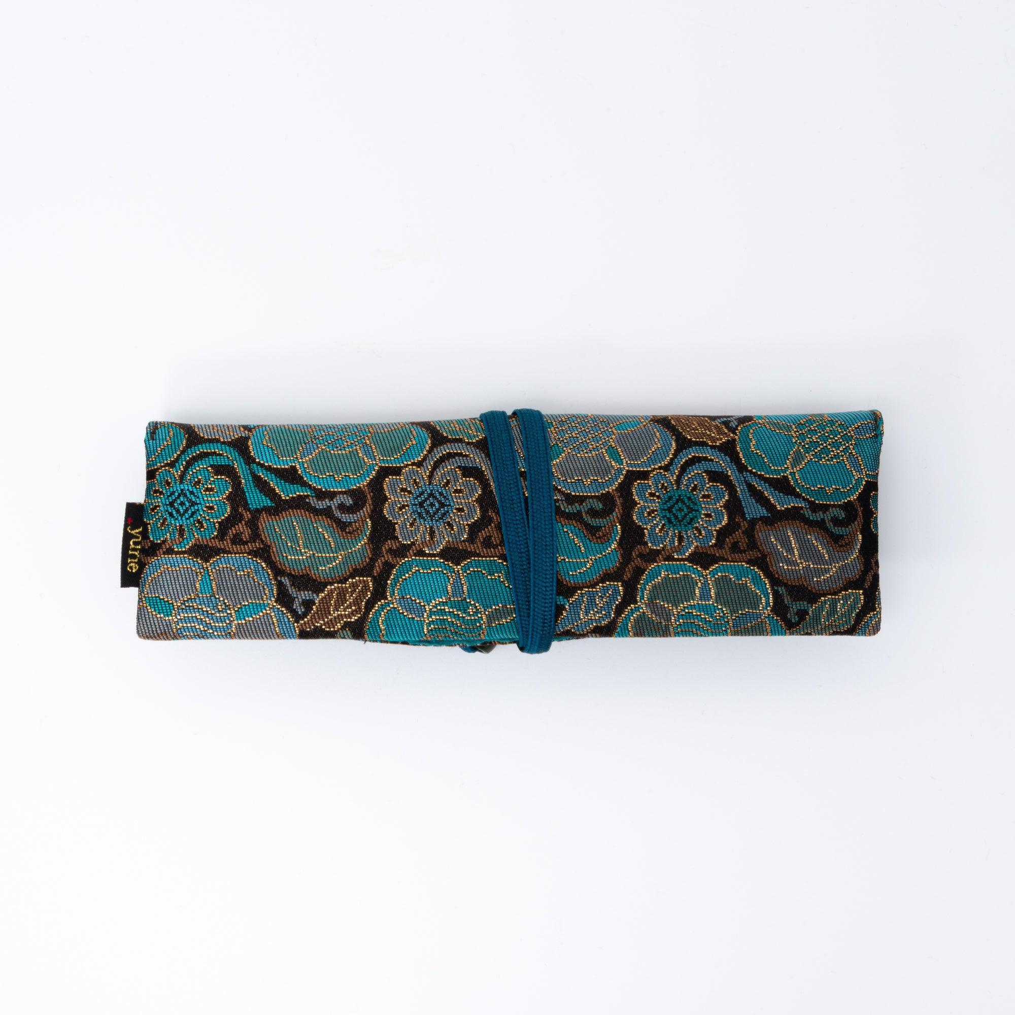 A neatly folded teal and gold Japanese fountain pen case of Nishijin silk, secured with a blue elastic cord on a white background.