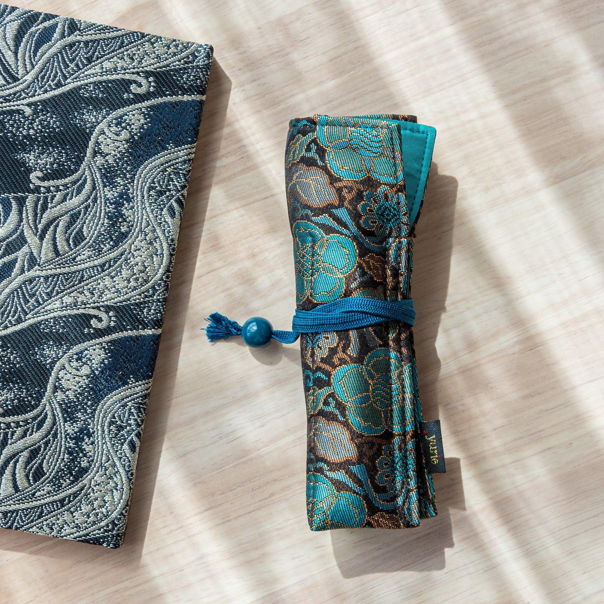 A rolled-up teal and gold Japanese fountain pen case made of Nishijin silk, secured with a blue cord, on a light wooden surface.