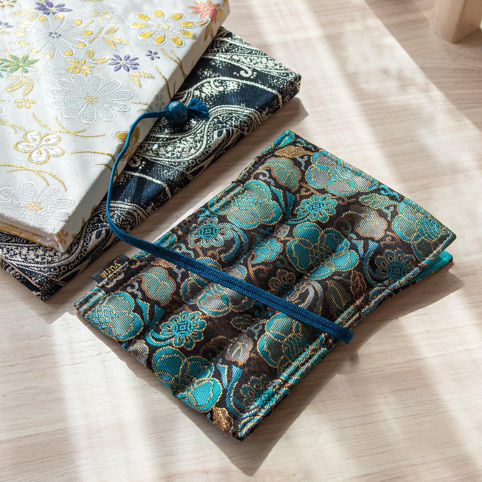 A teal and gold Japanese fountain pen case of Nishijin silk, placed next to white and navy patterned cases on a light wooden surface.
