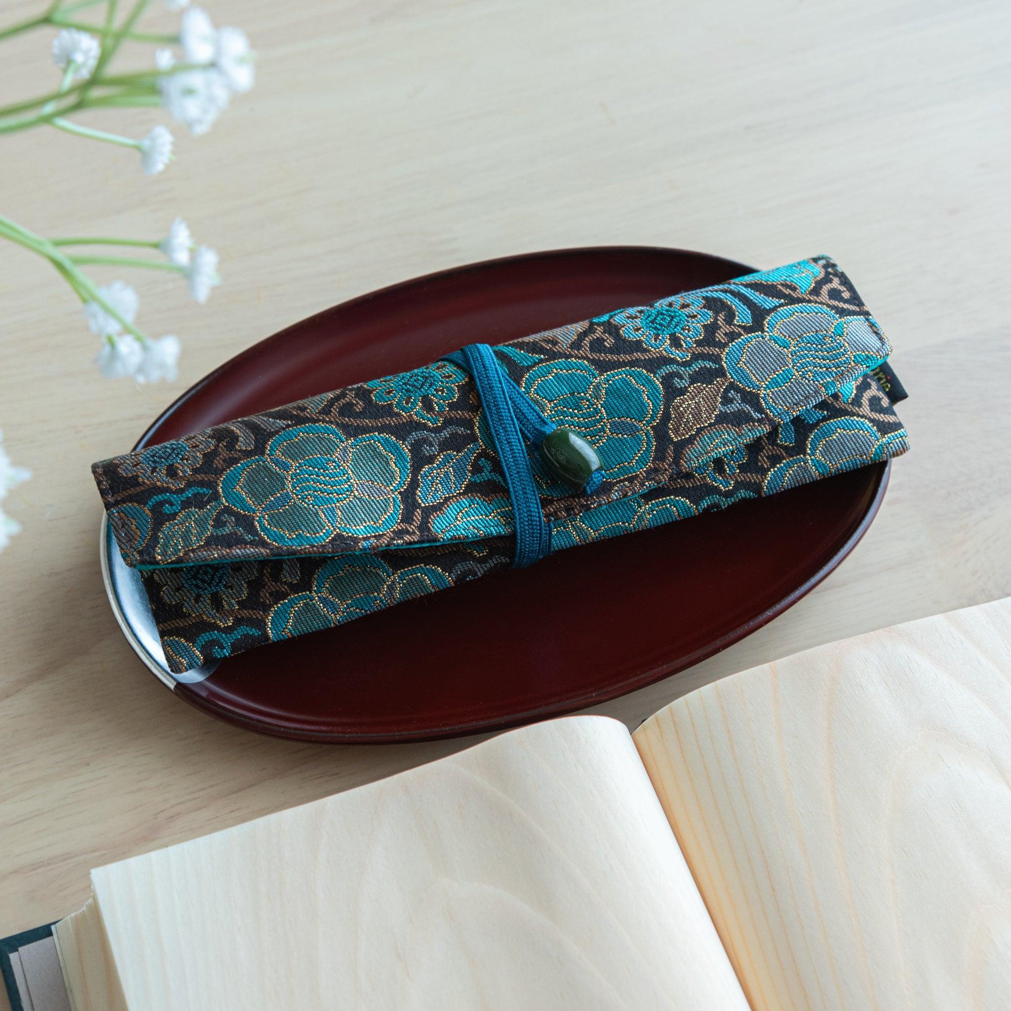 A folded teal and gold Japanese fountain pen case of Nishijin silk, tied neatly with a blue cord, resting on a red plate.