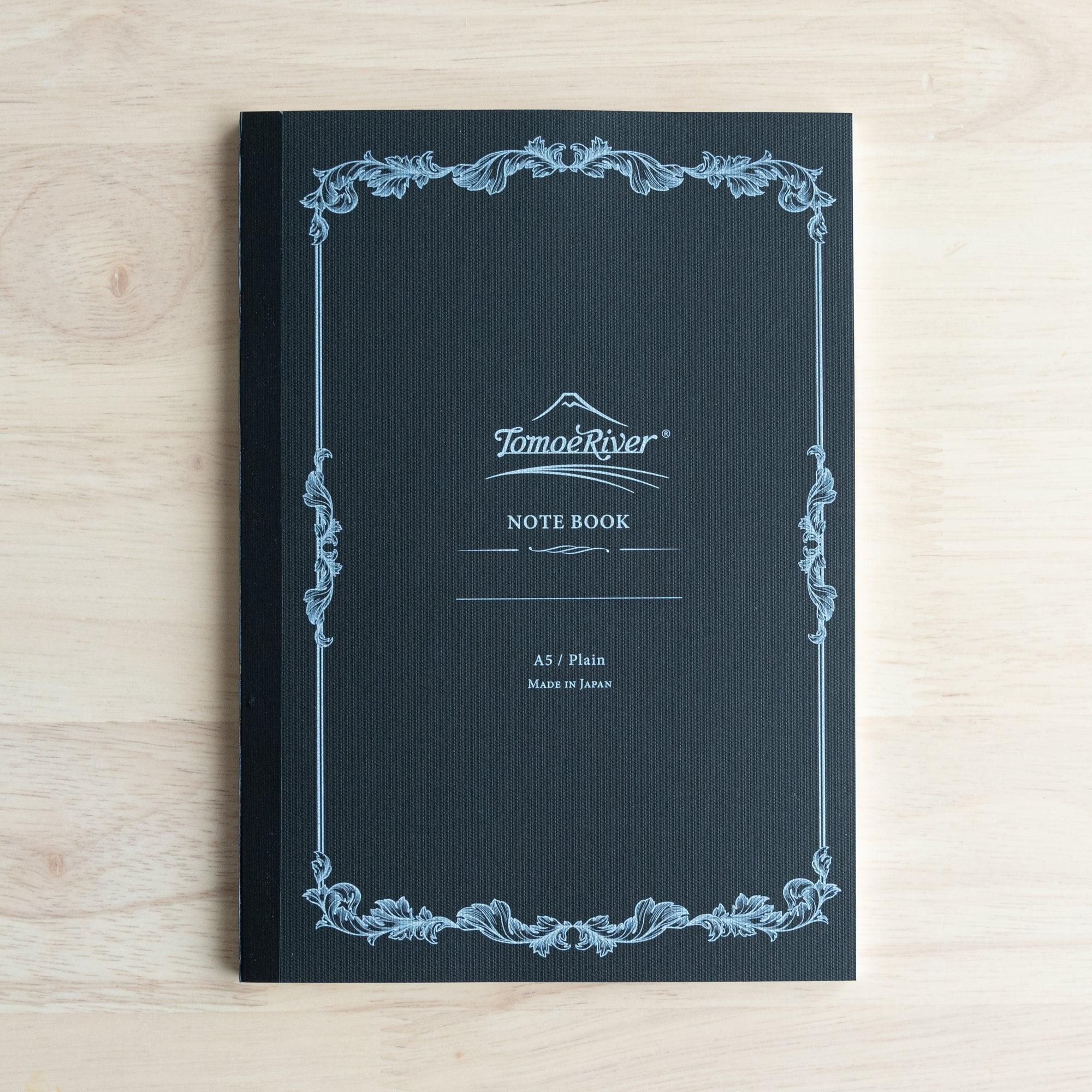A single black Tomoe River notebook with decorative borders placed on a wooden table, featuring the "A5 / Plain" label.