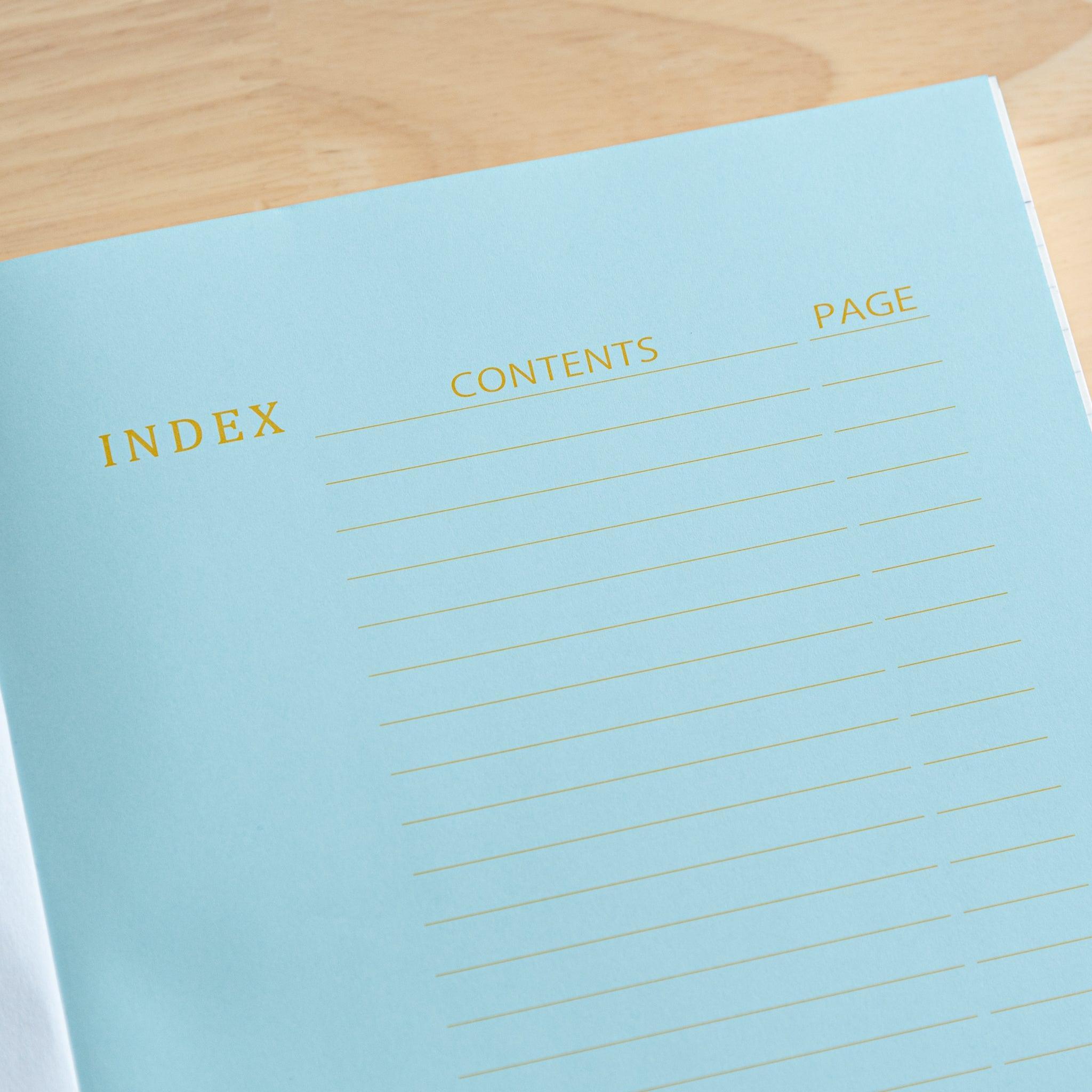An open Japanese notebook displaying a light blue index page with gold lettering and a black ribbon bookmark, showcasing fine Japanese stationery.