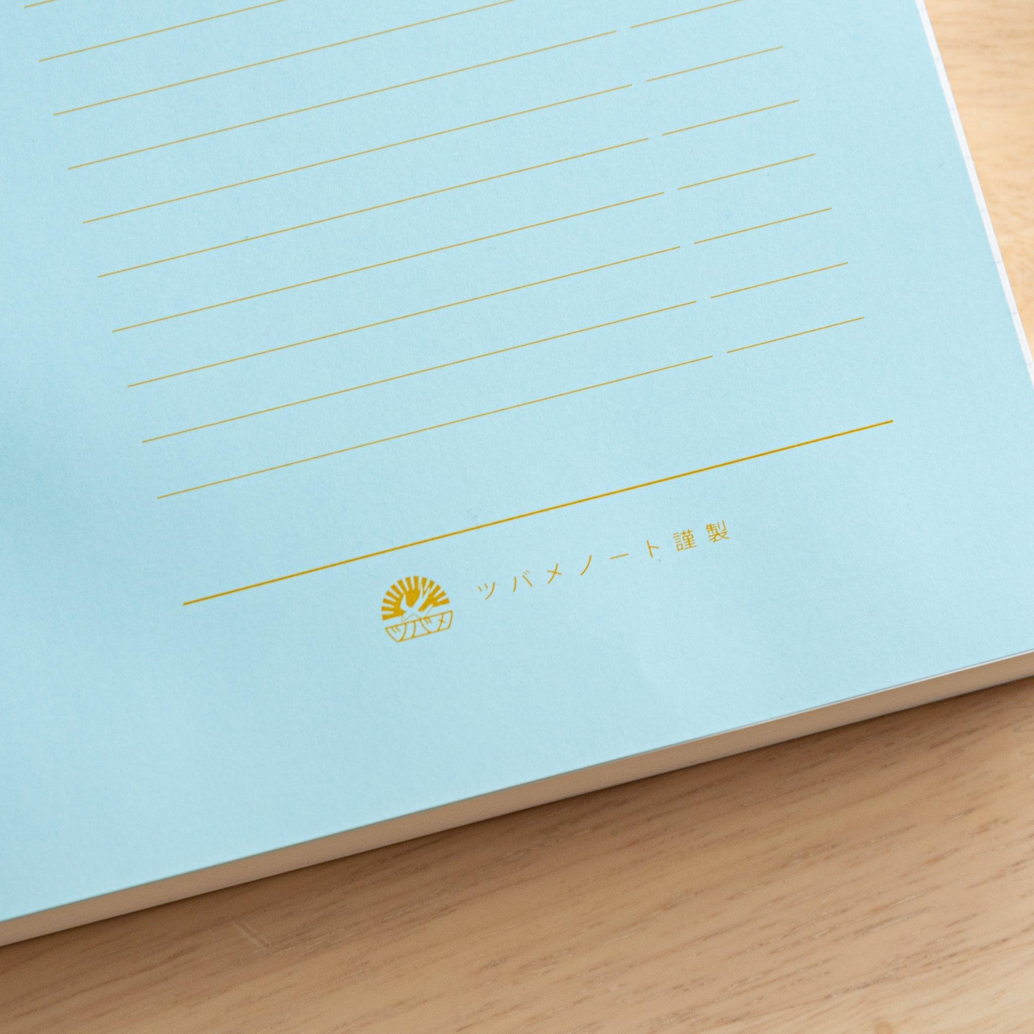 An open Japanese notebook displaying a light blue index page with gold lettering and a black ribbon bookmark, showcasing fine Japanese stationery.