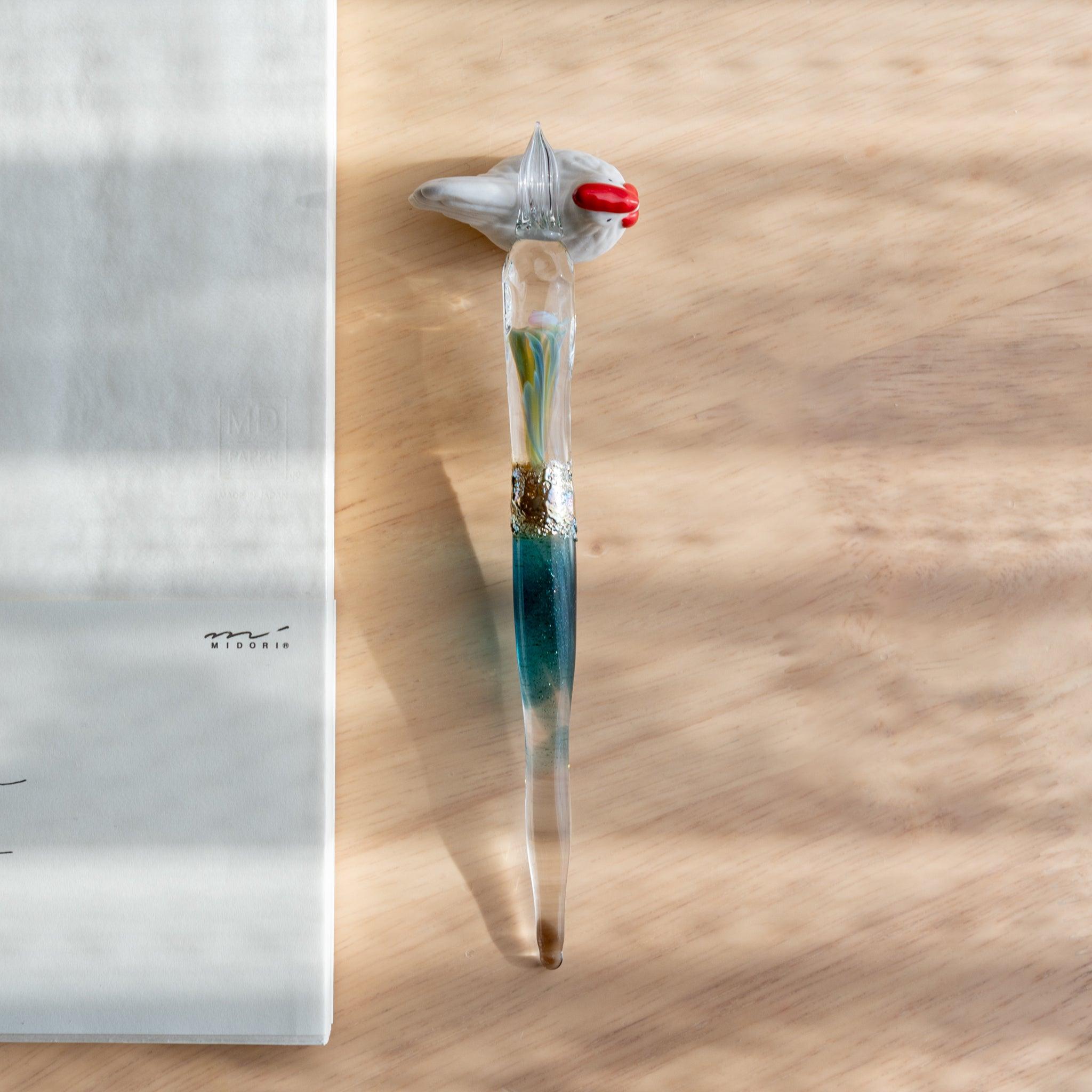 A Japanese glass dip pen with embedded opal and swirling colors rests on a bird-shaped ceramic holder. A white MD notebook lies nearby on a wooden desk.