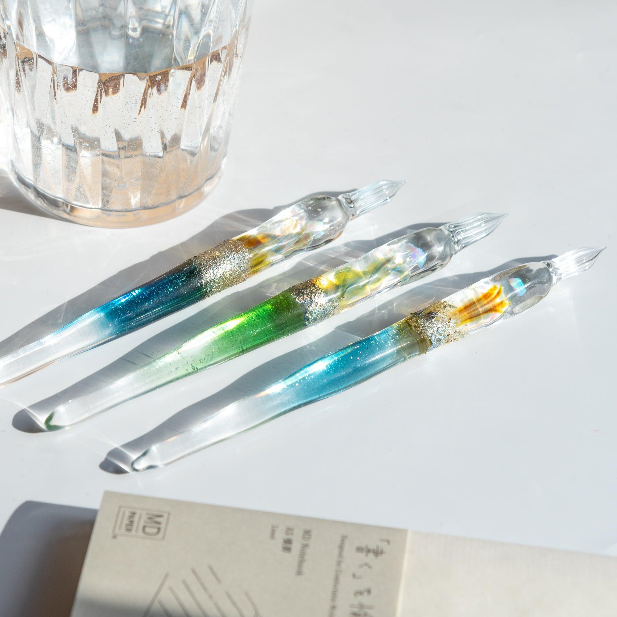 Three glass pens with suspended opals and silver accents, each with a unique color gradient, displayed in sunlight. Perfect for elegant writing.