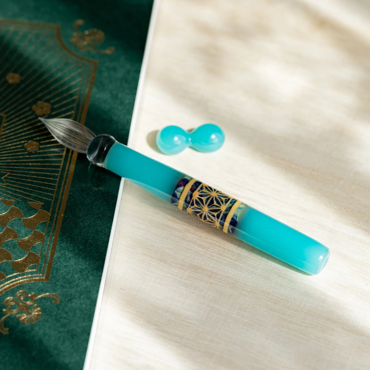 Turquoise Hemp Leaves glass dip pen with a patterned band, beside its two-sphere turquoise glass pen rest