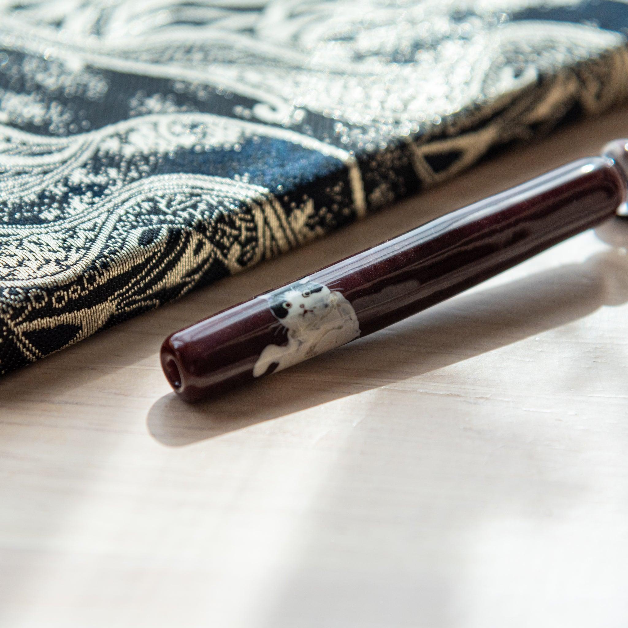 Dark red glass pen featuring a Tuxedo cat design, paired with a matching glass pen rest. Displayed on a wooden surface beside a Nishijin textile notebook.