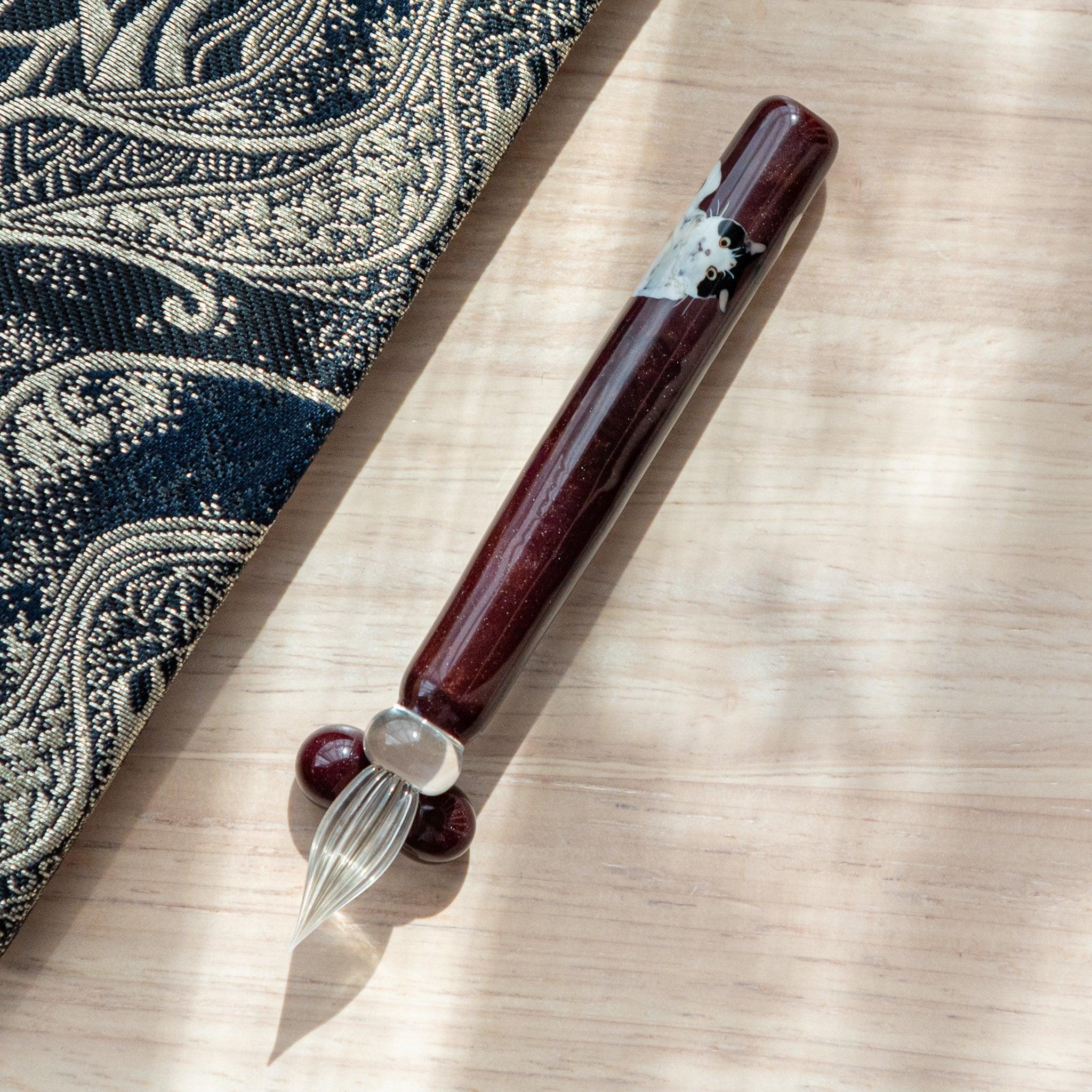 Dark red glass pen featuring a Tuxedo cat design, paired with a matching glass pen rest. Displayed on a wooden surface beside a Nishijin textile notebook.