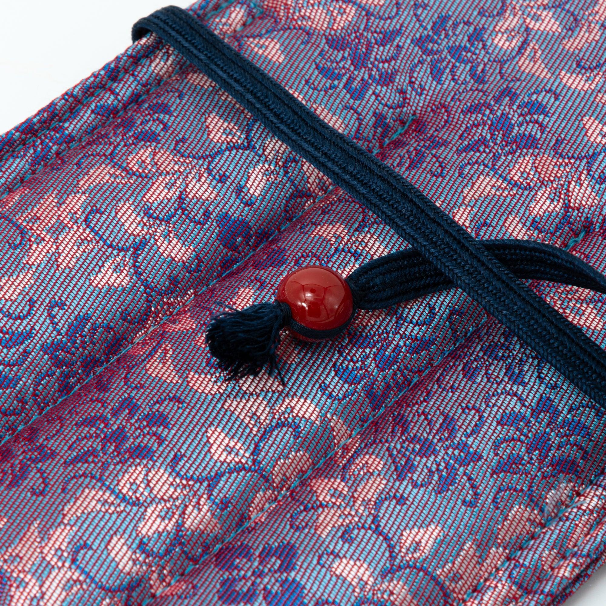 A folded Japanese fountain pen case in Nishijin silk with a floral pattern in pink and blue hues, secured with a navy cord and a red bead, displayed on a black tray.