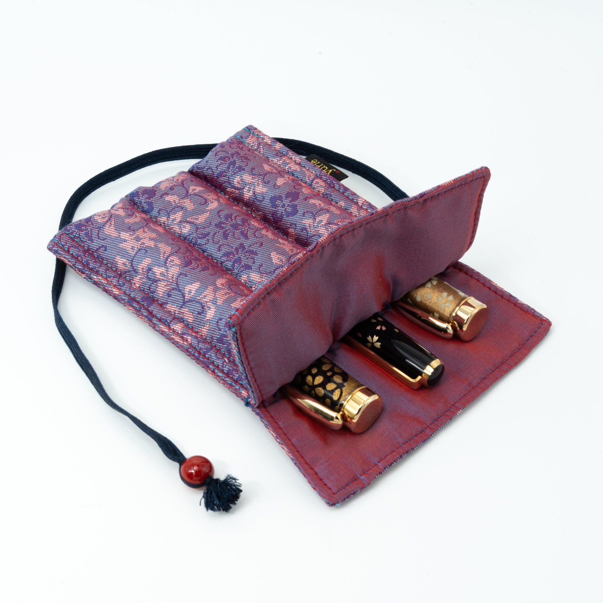 A Japanese fountain pen case in Nishijin silk with a blue and pink floral design, featuring a navy cord and red bead, placed on a black lacquer tray with a pink flower.