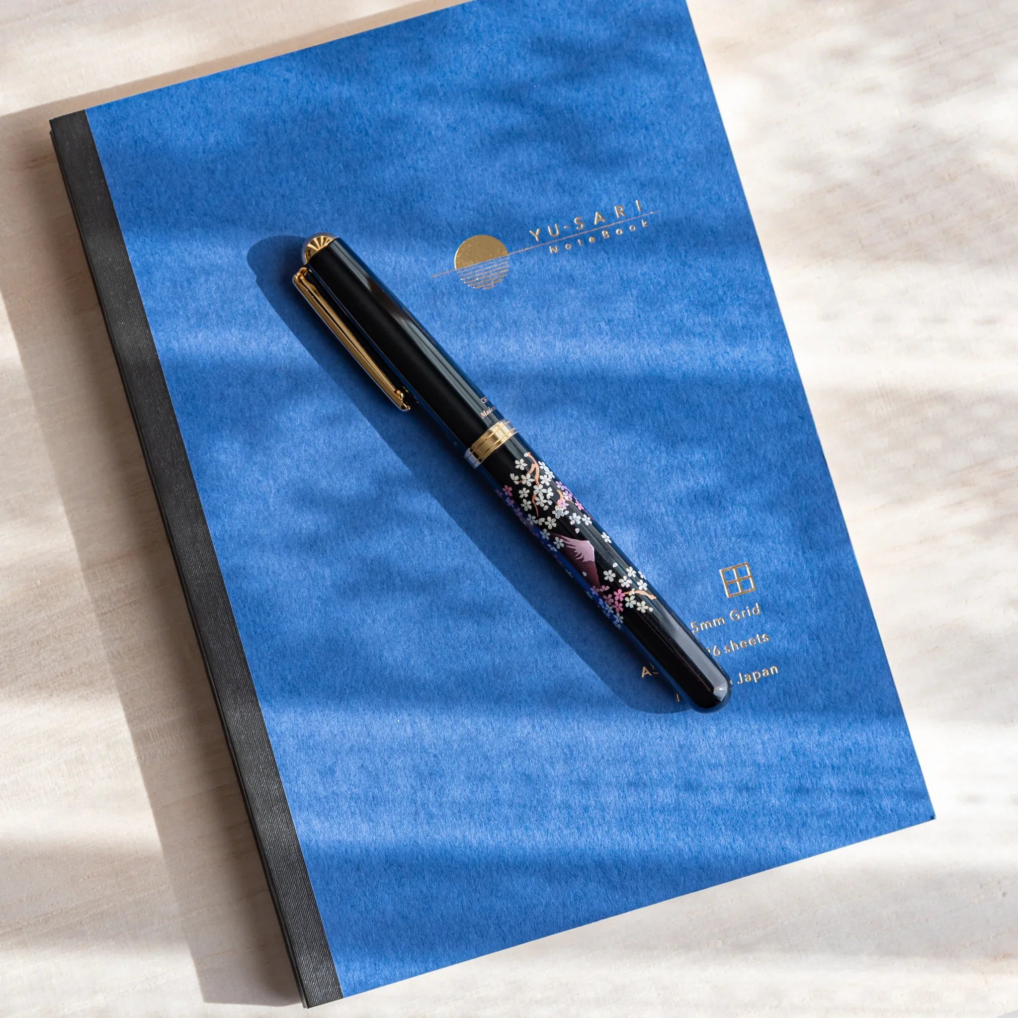 Black Japanese fountain pen with pink sakura and Mt. Fuji design resting on a blue notebook.