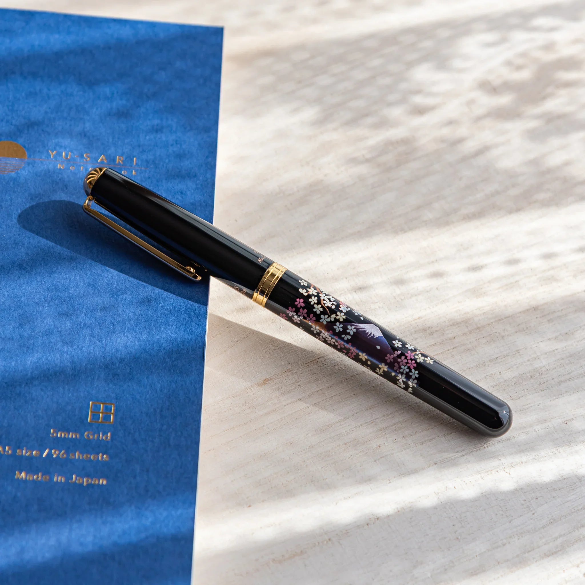 Black Japanese Fountain Pen with sakura and twilight Fuji design resting on a light desk beside a blue notebook.