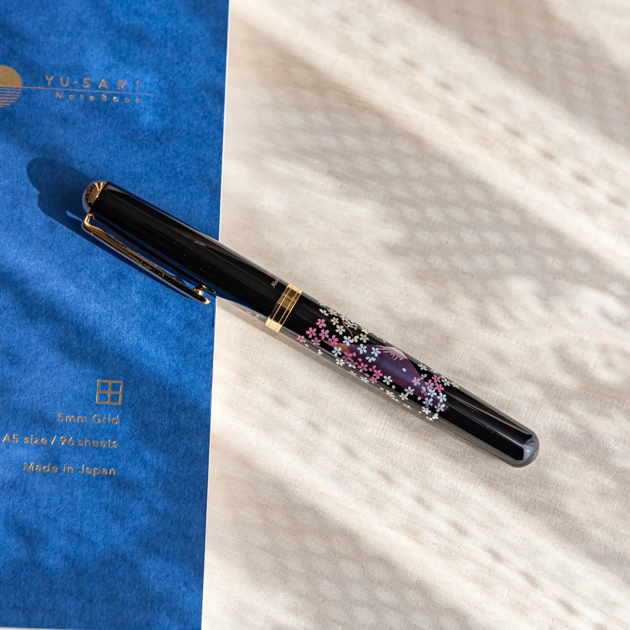 Japanese Fountain Pen with sakura and twilight Fuji design resting on a light desk beside a blue notebook.