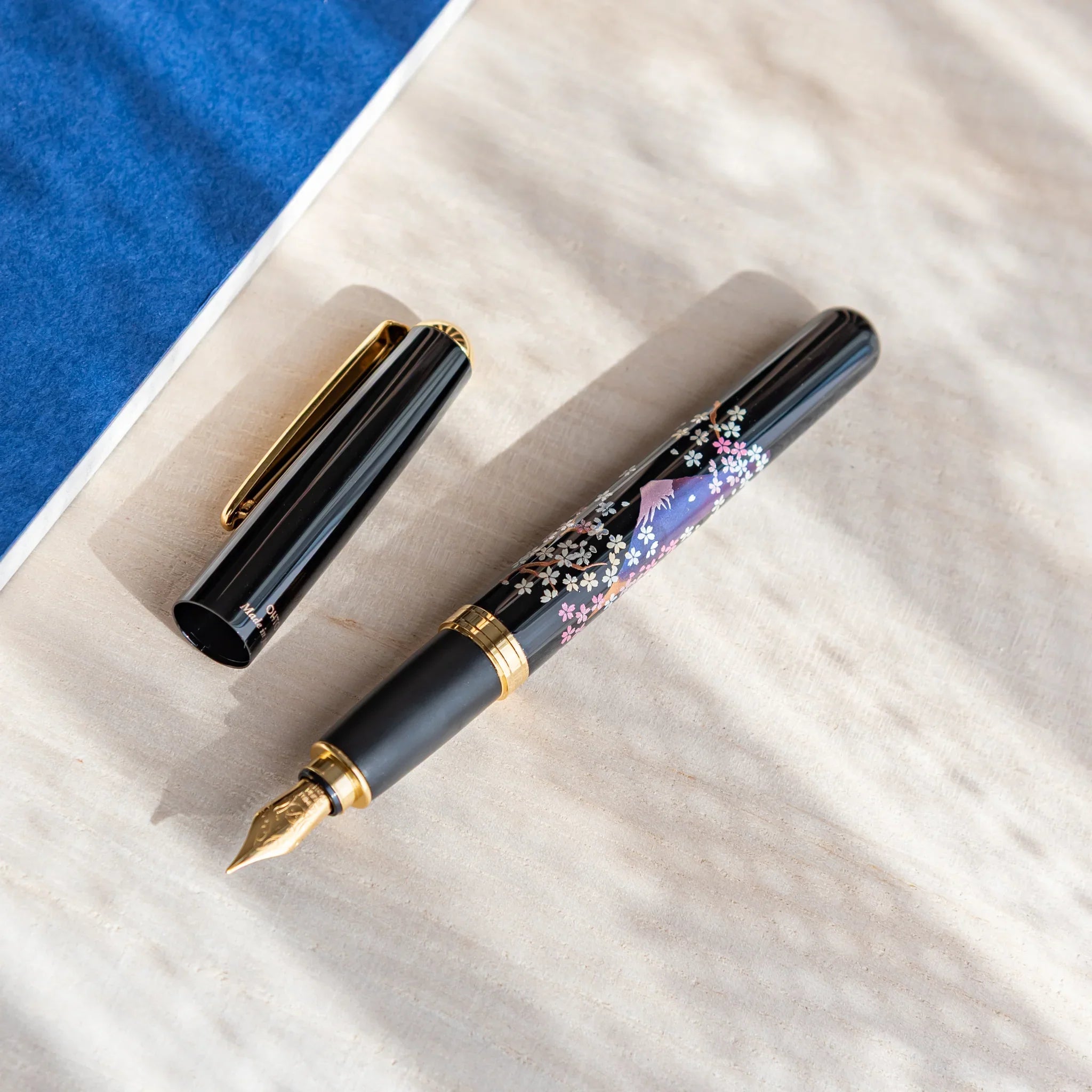 Uncapped Japanese Fountain Pen with twilight Fuji and sakura design resting on a light desk beside a blue notebook.