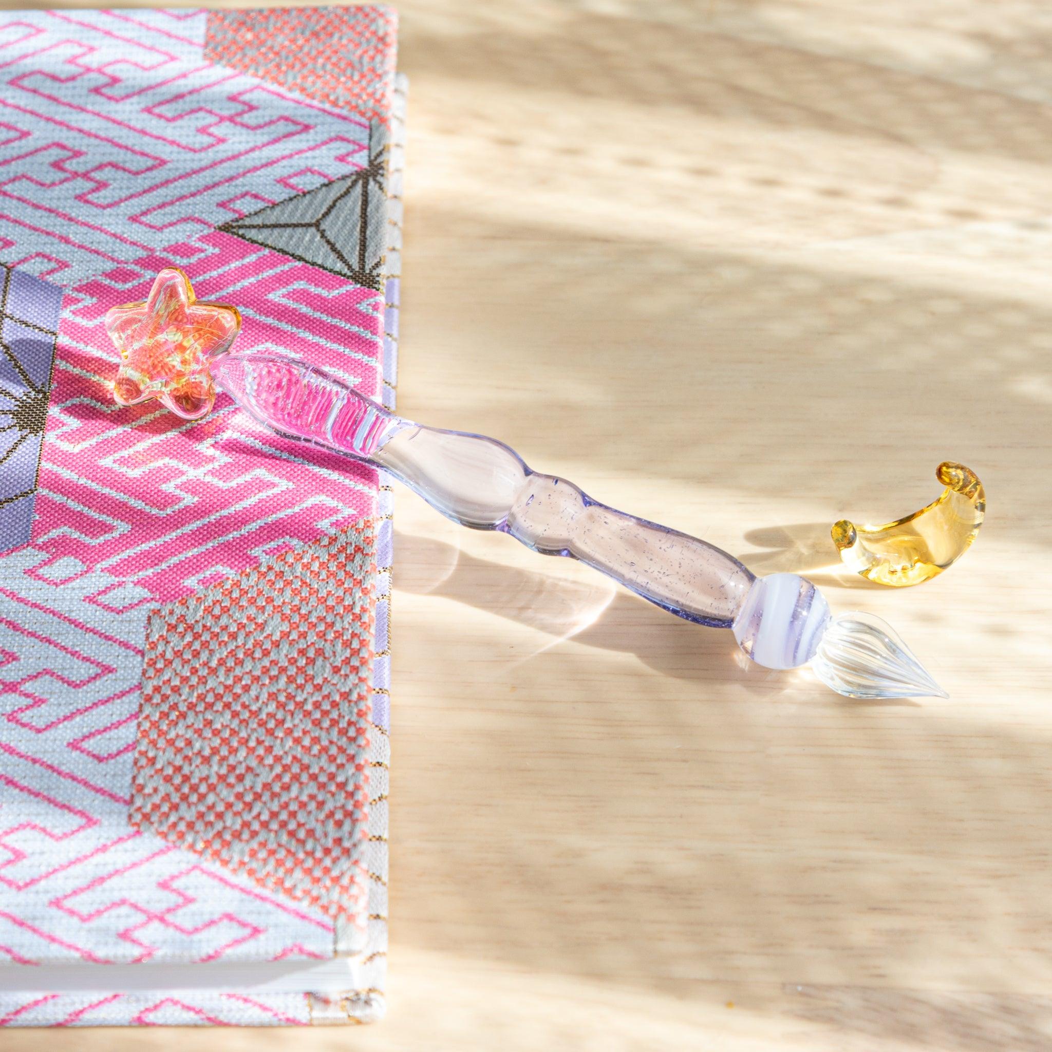 A lavender glass pen with a golden star top and a crescent moon pen rest lies on a wooden surface beside a Japanese notebook.