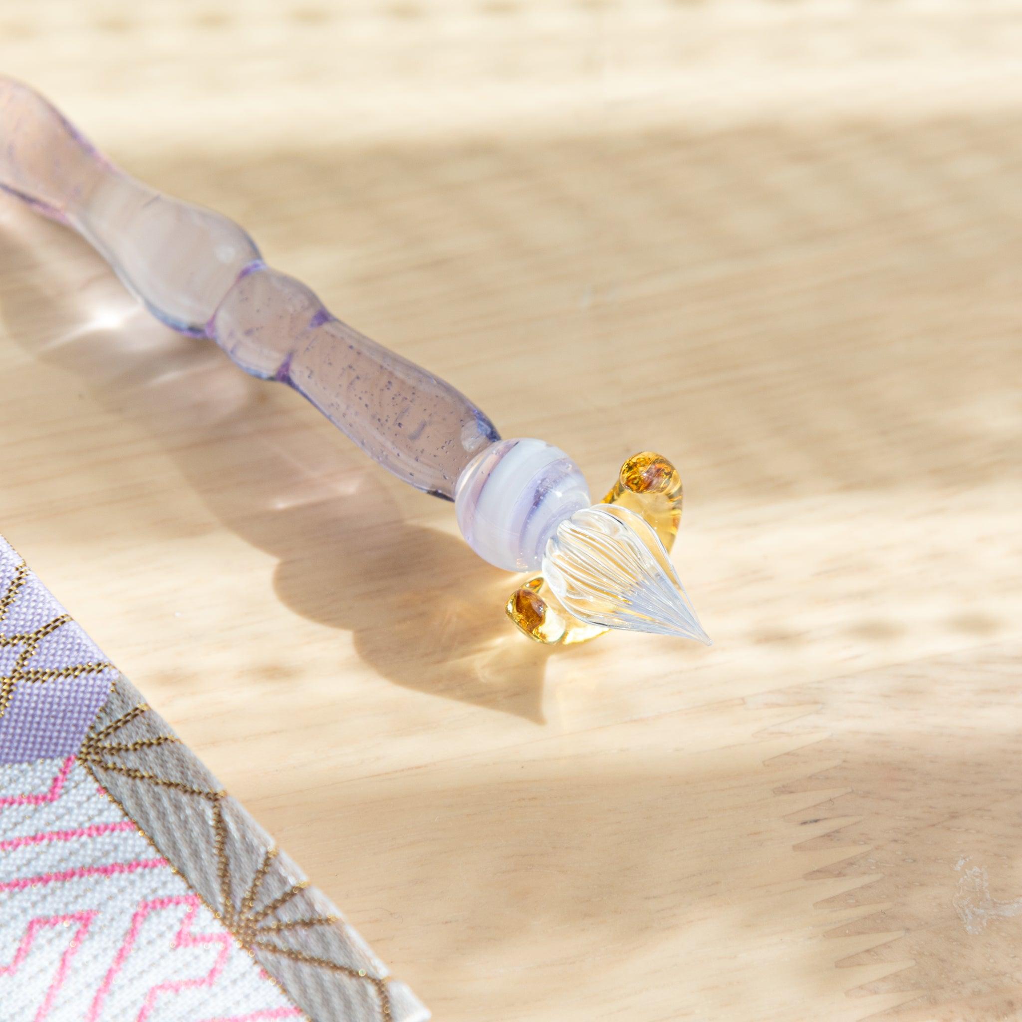 Close-up of the nib of a lavender glass pen with golden accents, resting on a wooden surface and reflecting warm sunlight.