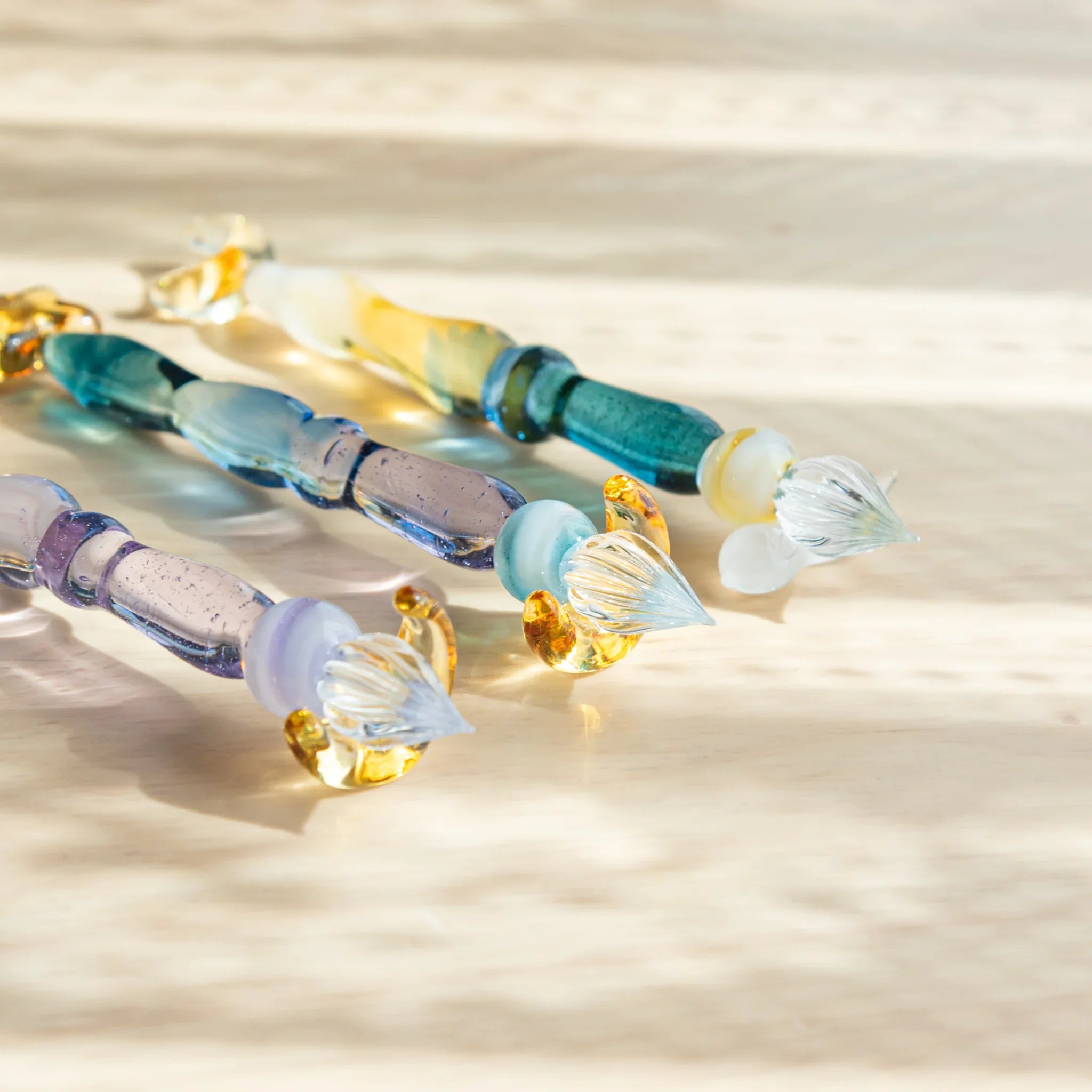 Three glass dip pens with floral glass nibs and amber accents on light wood showing barrel colors in teal, blue, and lavender