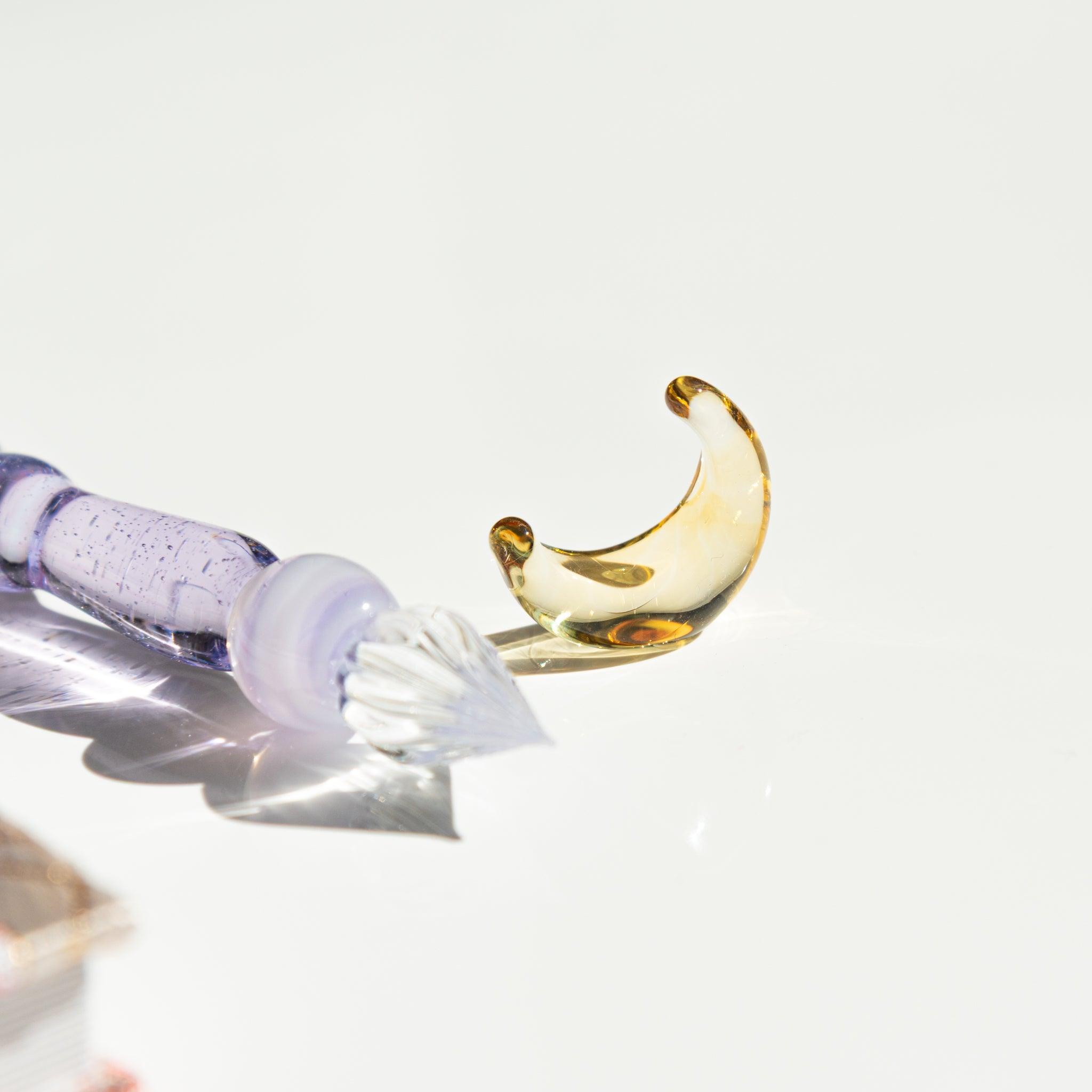Close-up of the nib of a lavender glass pen with a detached golden crescent moon accessory beside it, both reflecting light elegantly.