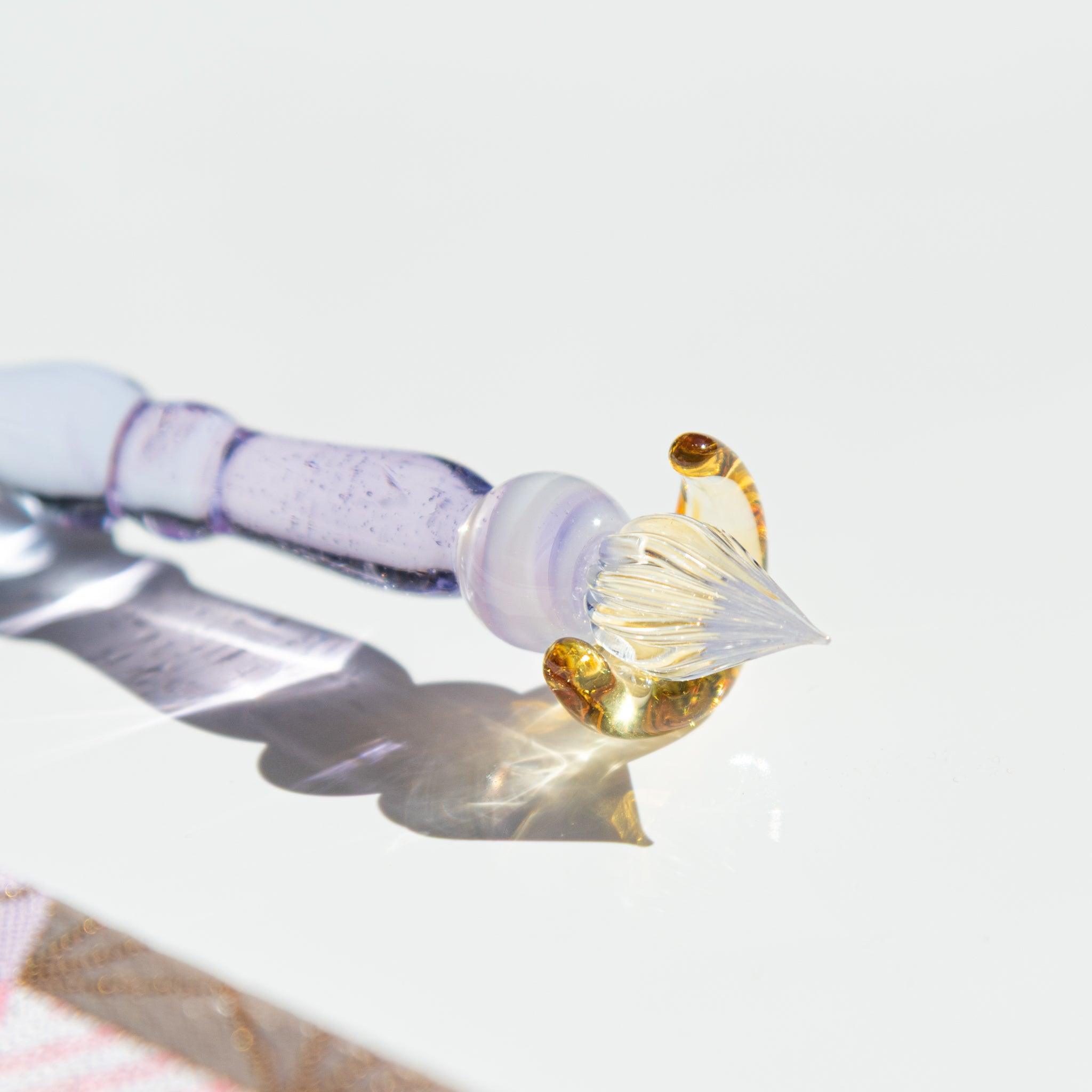 Close-up of the nib of a lavender glass pen with a golden crescent moon pen rest, reflecting light on a glossy white surface.