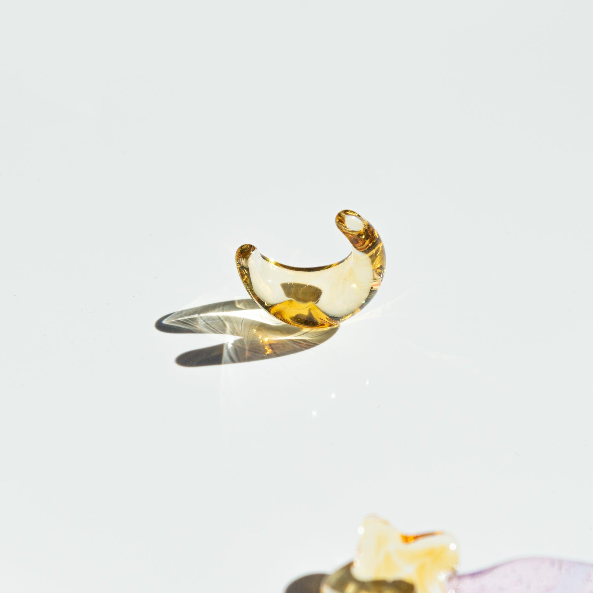 A golden crescent moon-shaped glass pen rest lies on a reflective white surface, casting a delicate shadow in the bright light.