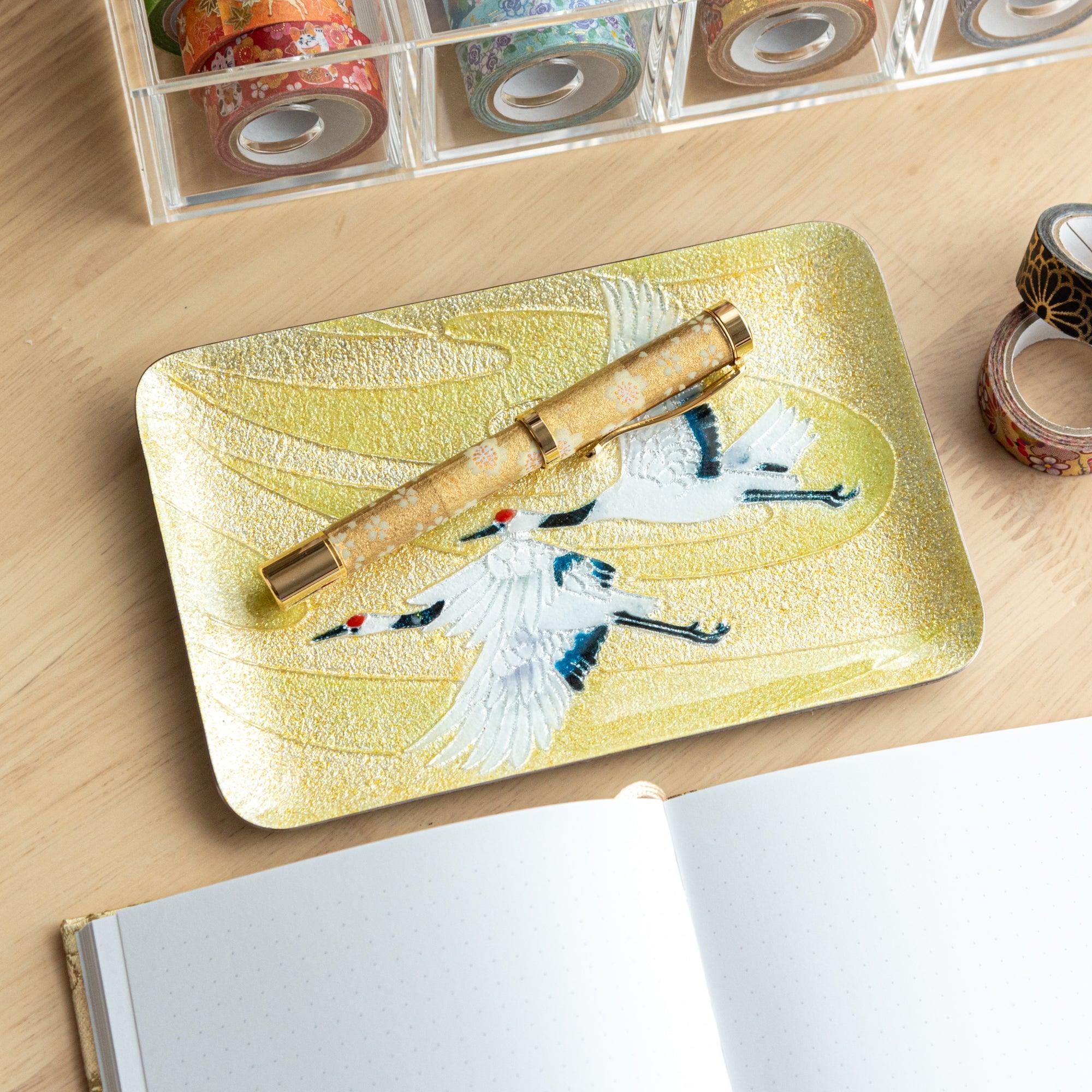 A gold-patterned fountain pen resting on a tray adorned with white cranes and a shimmering gold background, highlighting Japanese design.