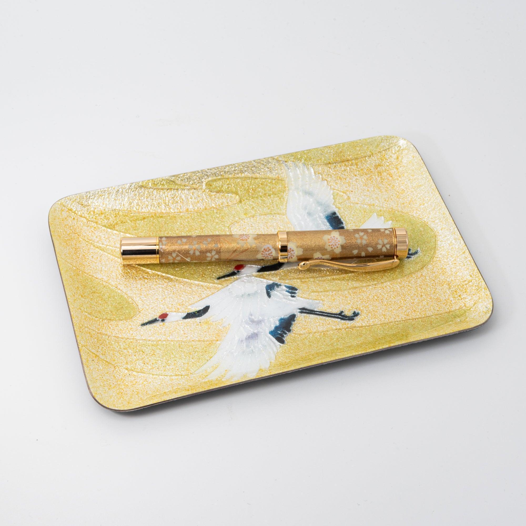 A close-up of a gold fountain pen on a decorative tray featuring two white cranes, symbolizing elegance in Japanese stationery.