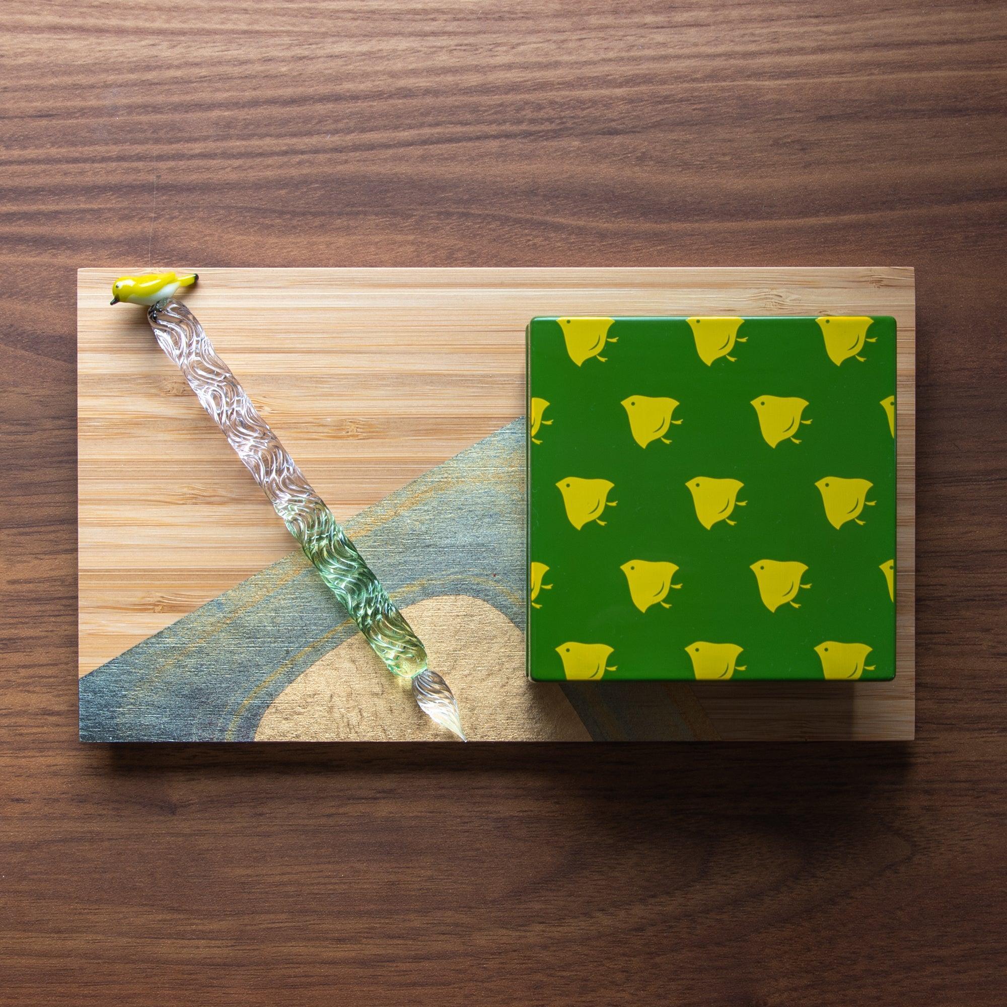 Green Japanese lacquer stationery box with yellow bird patterns, paired with a matching green glass pen on a decorative wooden board, creating a playful setup.