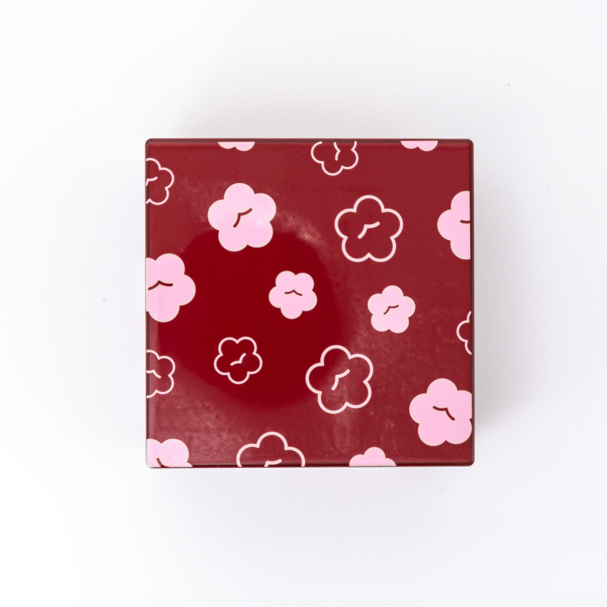 Top view of a red Japanese lacquer stationery box with pink plum blossom patterns, showcasing its decorative floral design and glossy finish on a white surface.