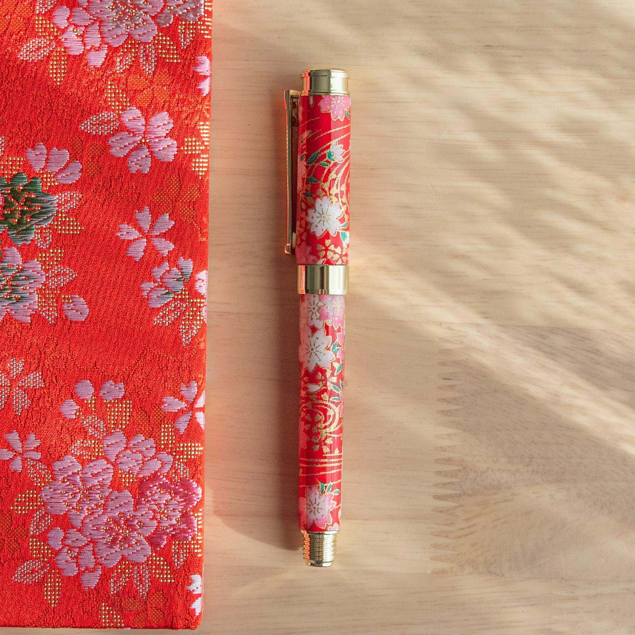 A red floral Japanese stationery fountain pen placed on a table next to matching fabric under sunlight.