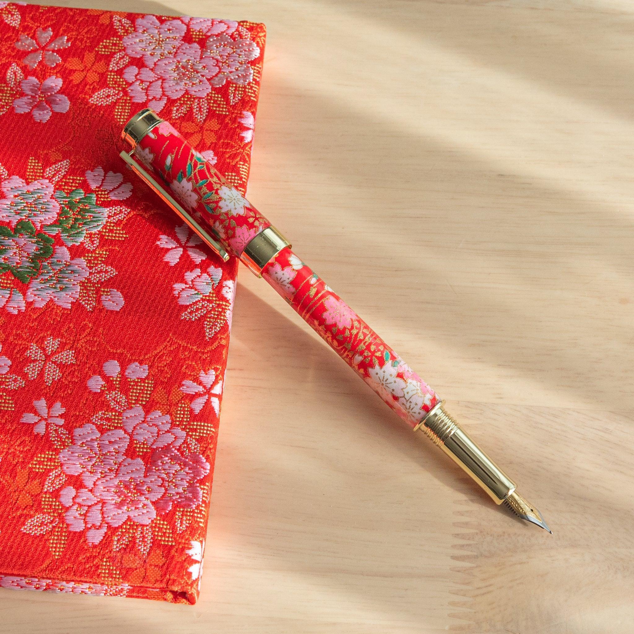 A red floral Japanese stationery fountain pen with its nib exposed, placed beside matching fabric under sunlight.