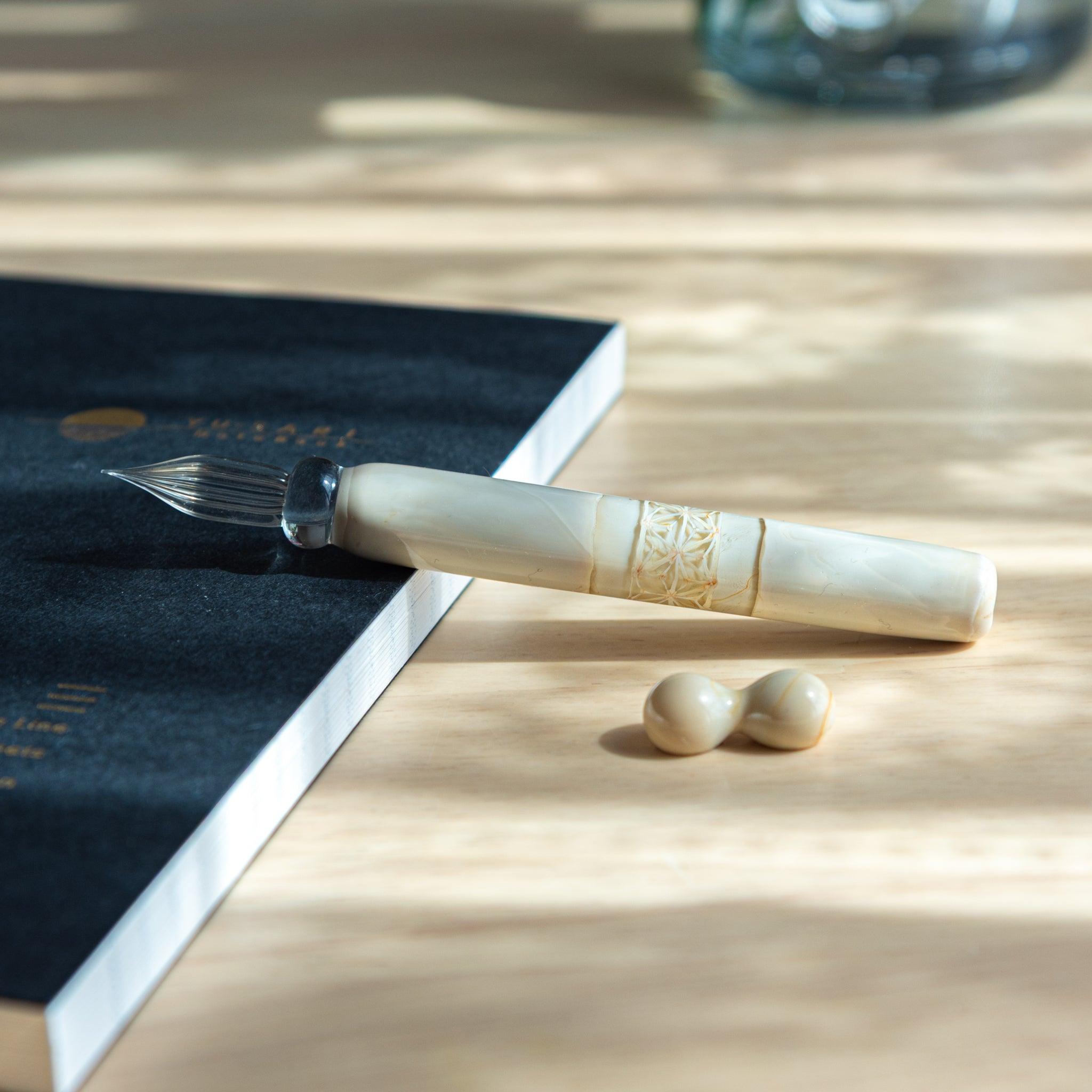 A Japanese glass dip pen with a carved ivory-colored handle rests on a notebook, bathed in soft natural light on a wooden surface.