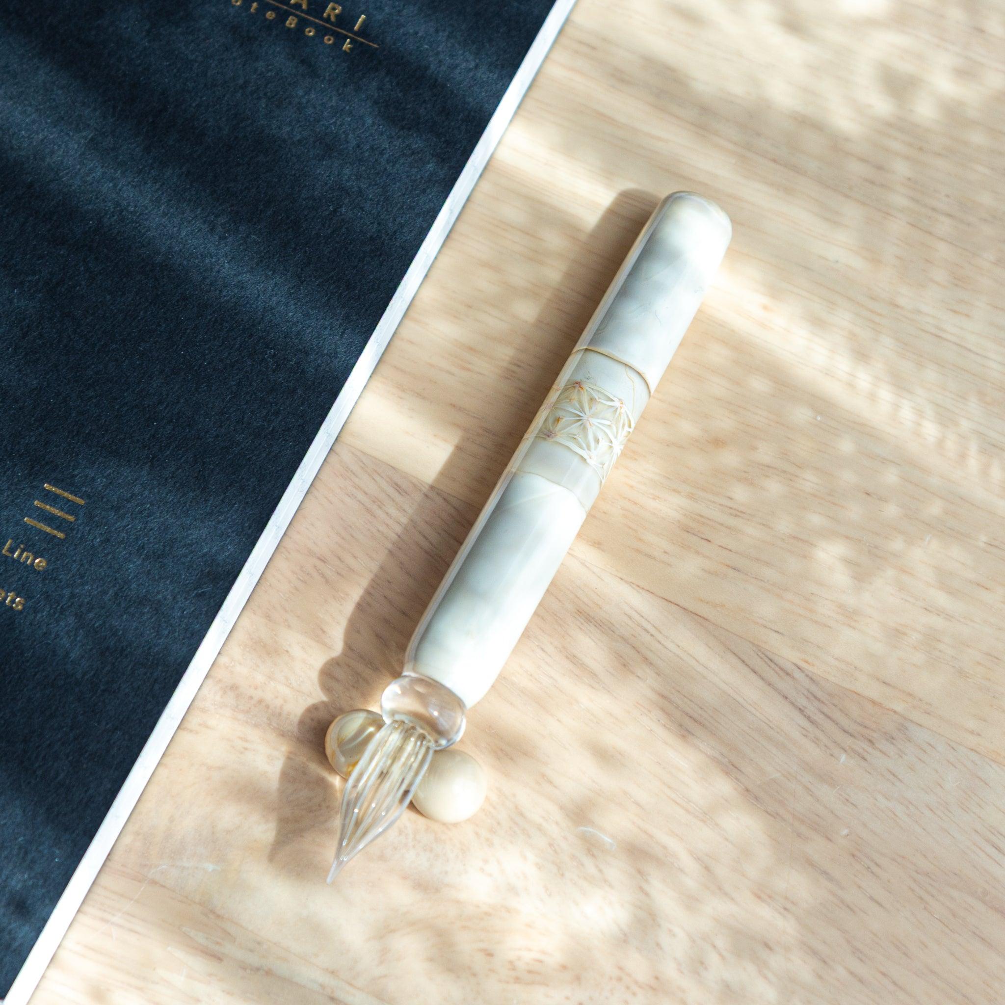 A Japanese glass dip pen with a creamy marbled handle and intricate carvings lies on a wooden surface next to a dark notebook.