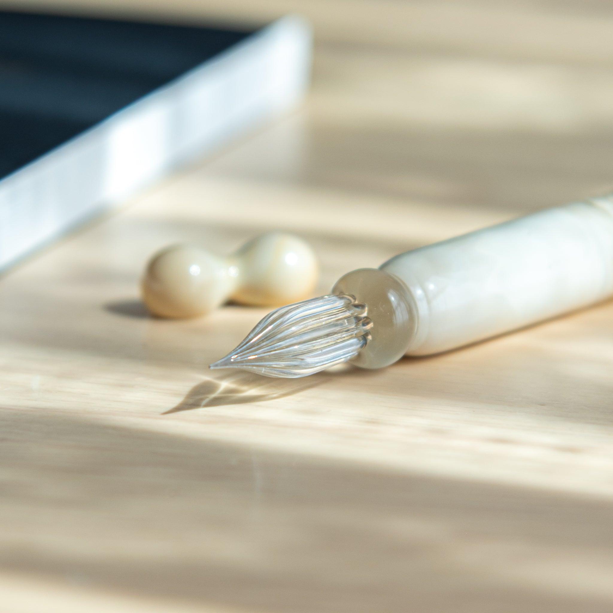 Close-up of a Japanese glass dip pen's twisted clear nib and ivory-toned handle, placed on a wooden surface in soft natural light.