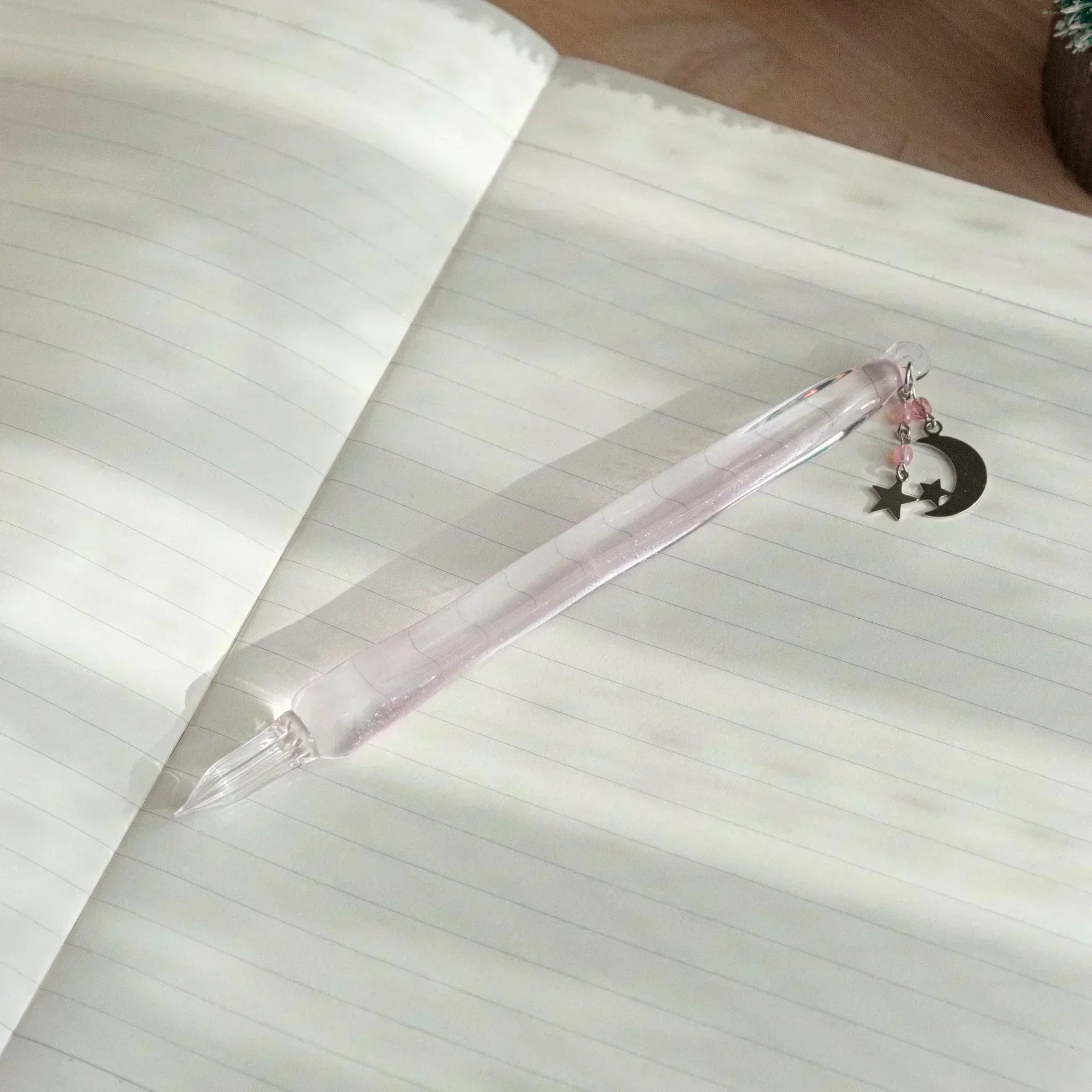 Glass dip pen with a hint of pink, moon and star charm, and pink bead decorations, resting diagonally on an open notebook with lined pages.