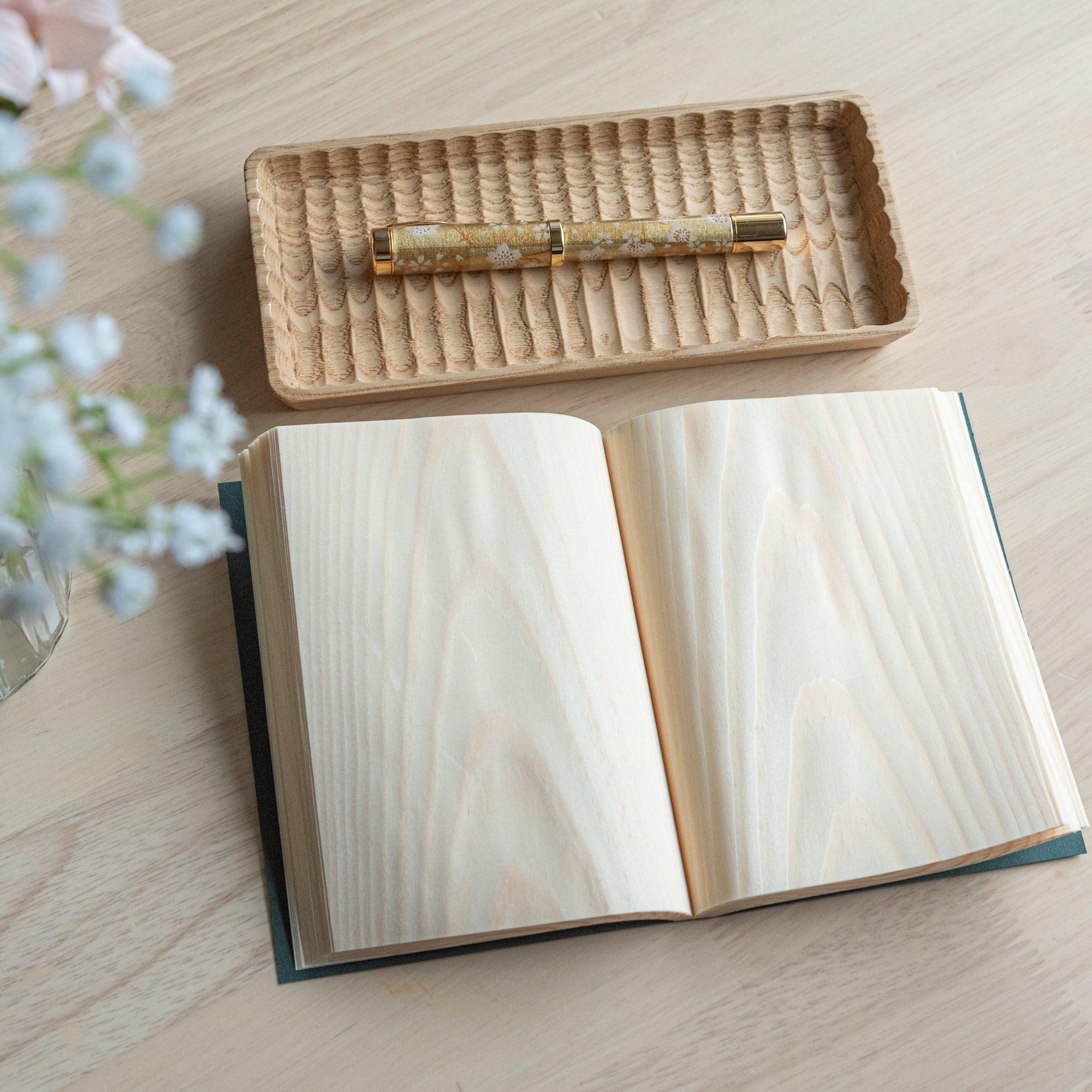 Open Japanese notebook with wooden grain pages, a gold floral pen on a wooden tray, and a vase with flowers, showcasing Japanese stationery.