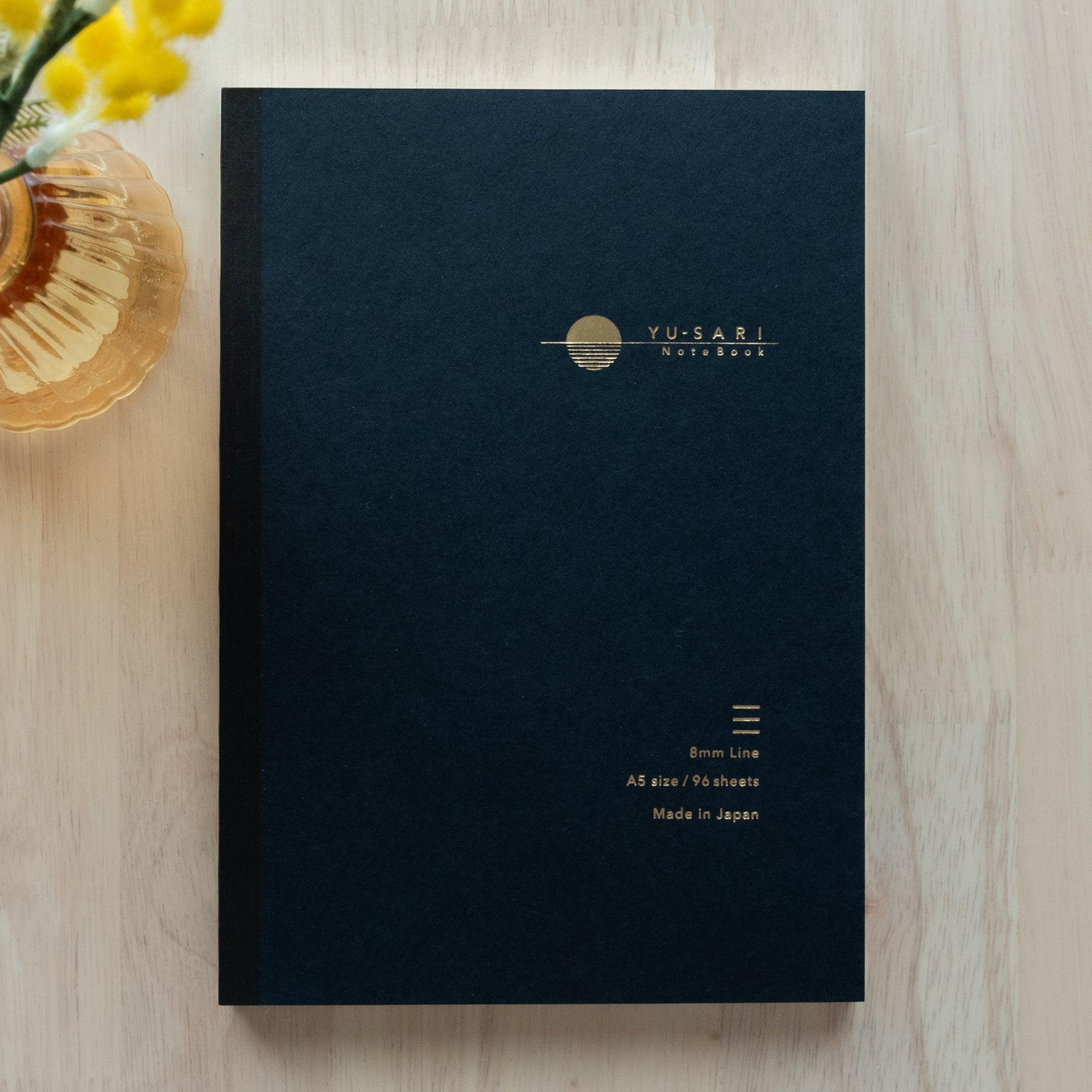 black notebook cover with gold-embossed "YU-SARI Notebook" logo, a minimal sun design, and specifications on a light wooden table.