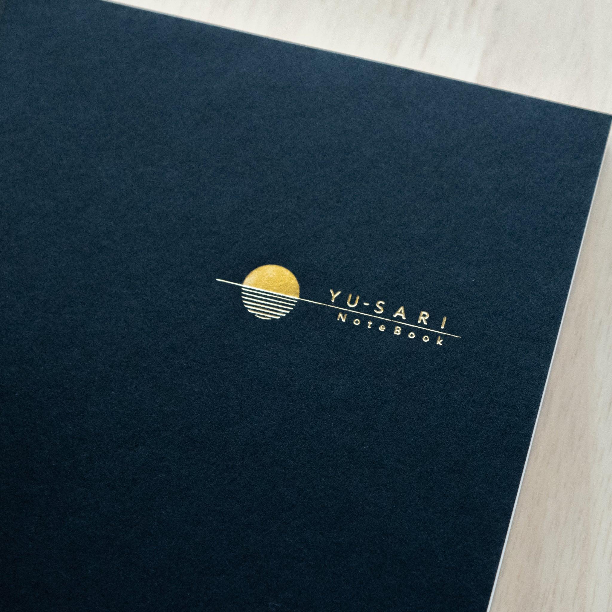 Close-up of the "YU-SARI Notebook" logo on a black cover, featuring a gold-embossed minimal sun design with horizontal lines.