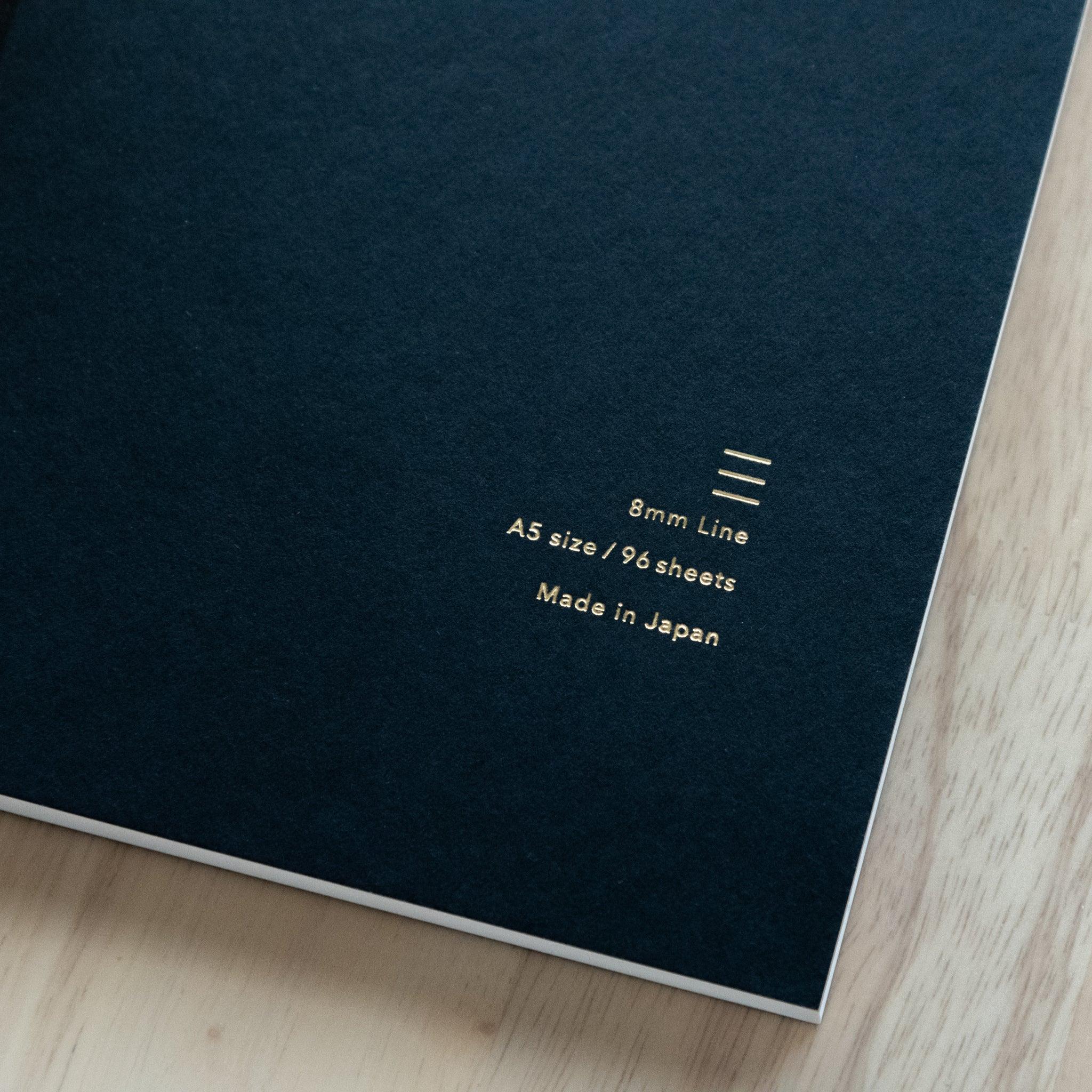 black notebook cover with gold-embossed text, reading "8mm Line, A5 size / 96 sheets, Made in Japan" on a wooden surface.