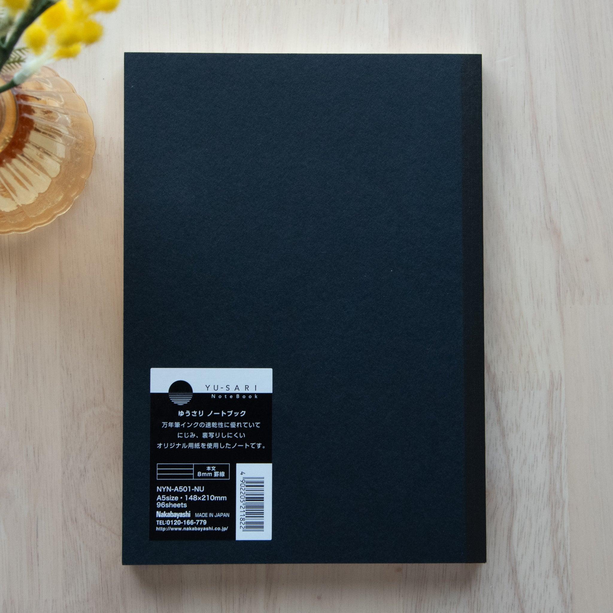 Back cover of a black notebook with a white and black label featuring product details in Japanese and English on a light wooden table.