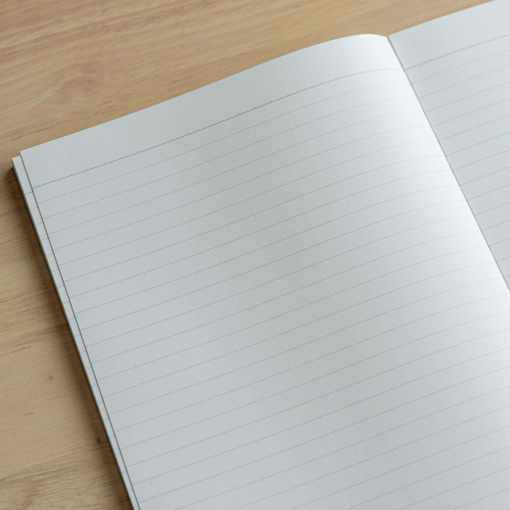 Close-up of an open notebook with lined white pages, showing subtle stitching along the spine on a light wooden table.
