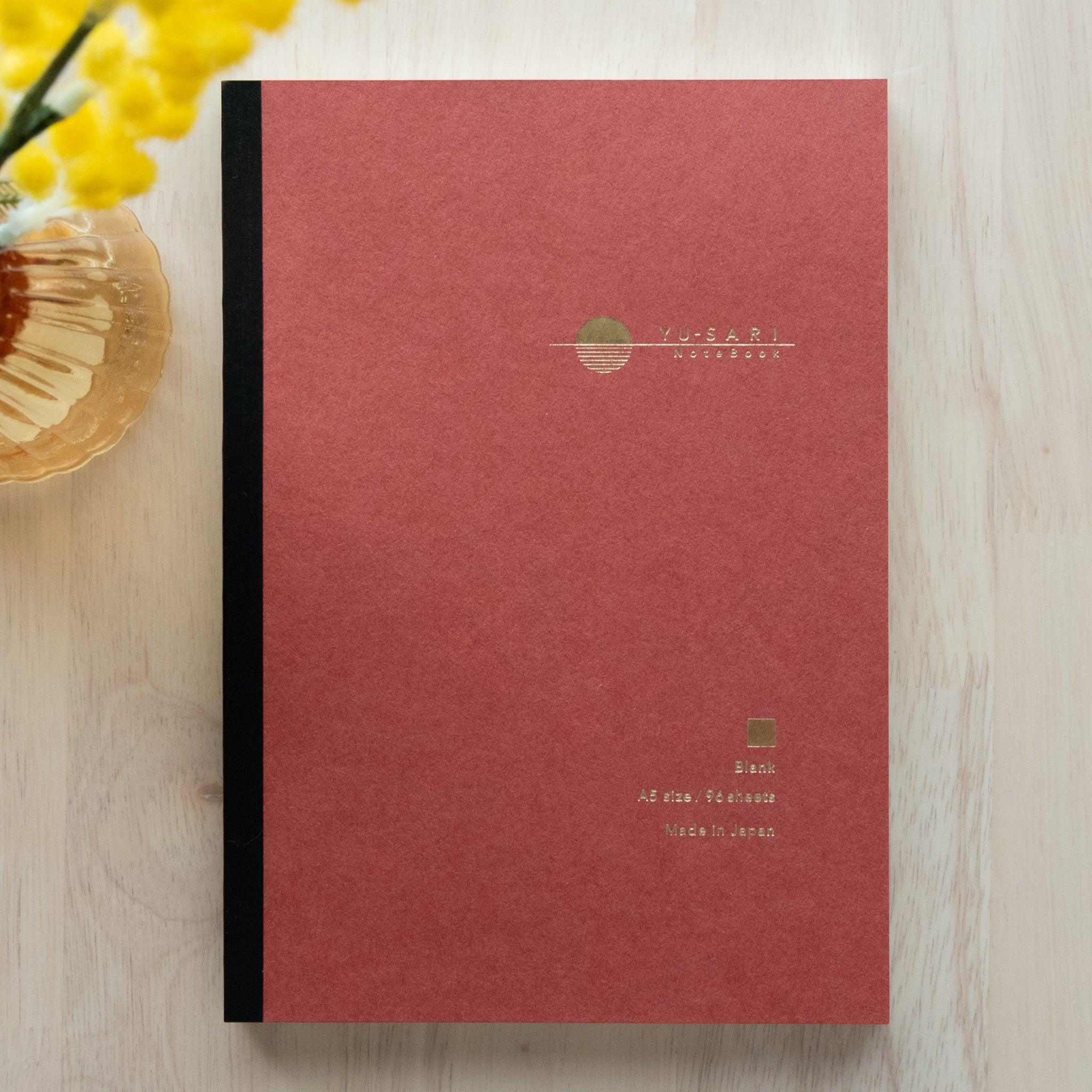 An open YU-SARI notebook revealing its smooth blank pages, ideal for writing or drawing, displayed on a wooden surface under soft light.