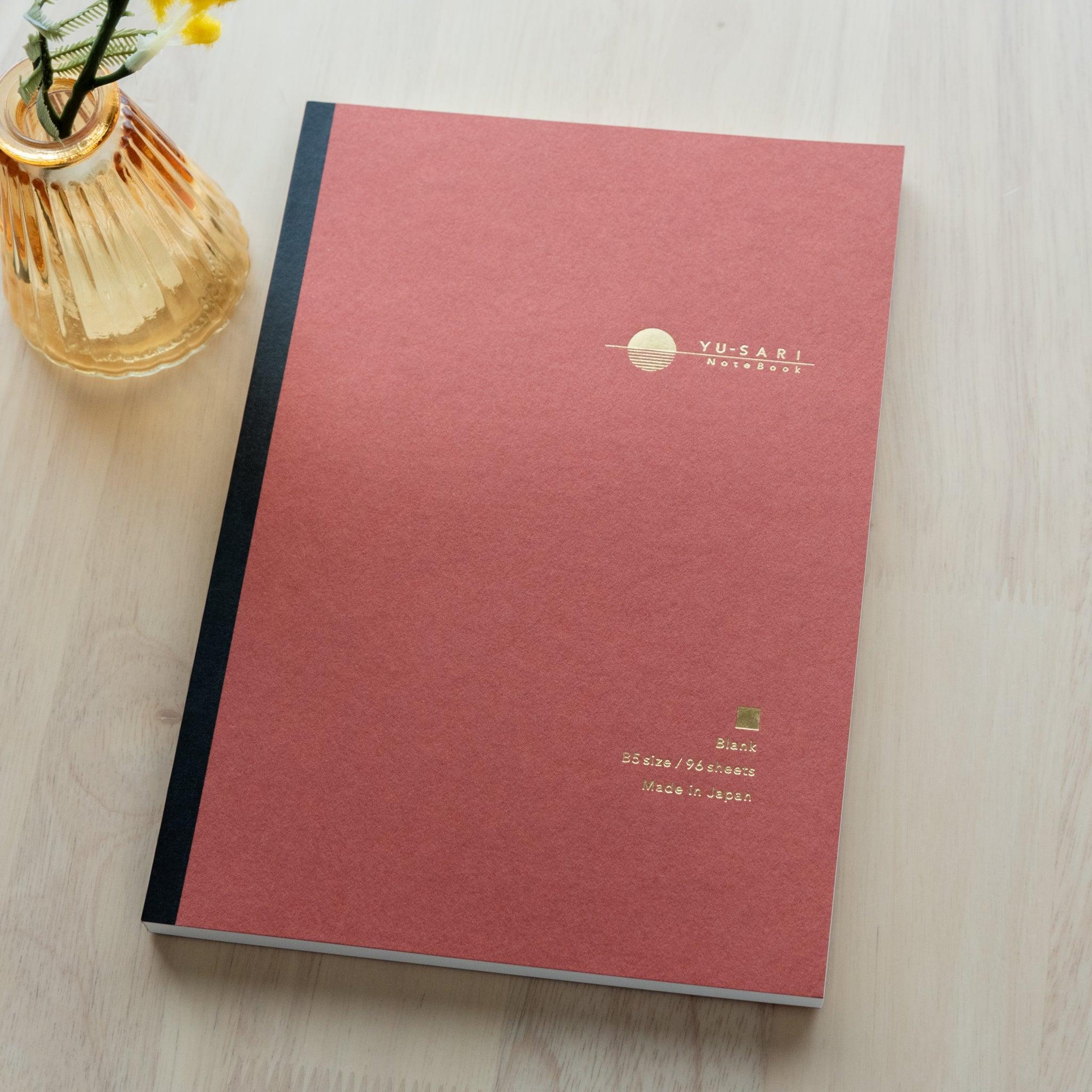 An open YU-SARI notebook revealing its smooth blank pages, ideal for writing or drawing, displayed on a wooden surface under soft light.