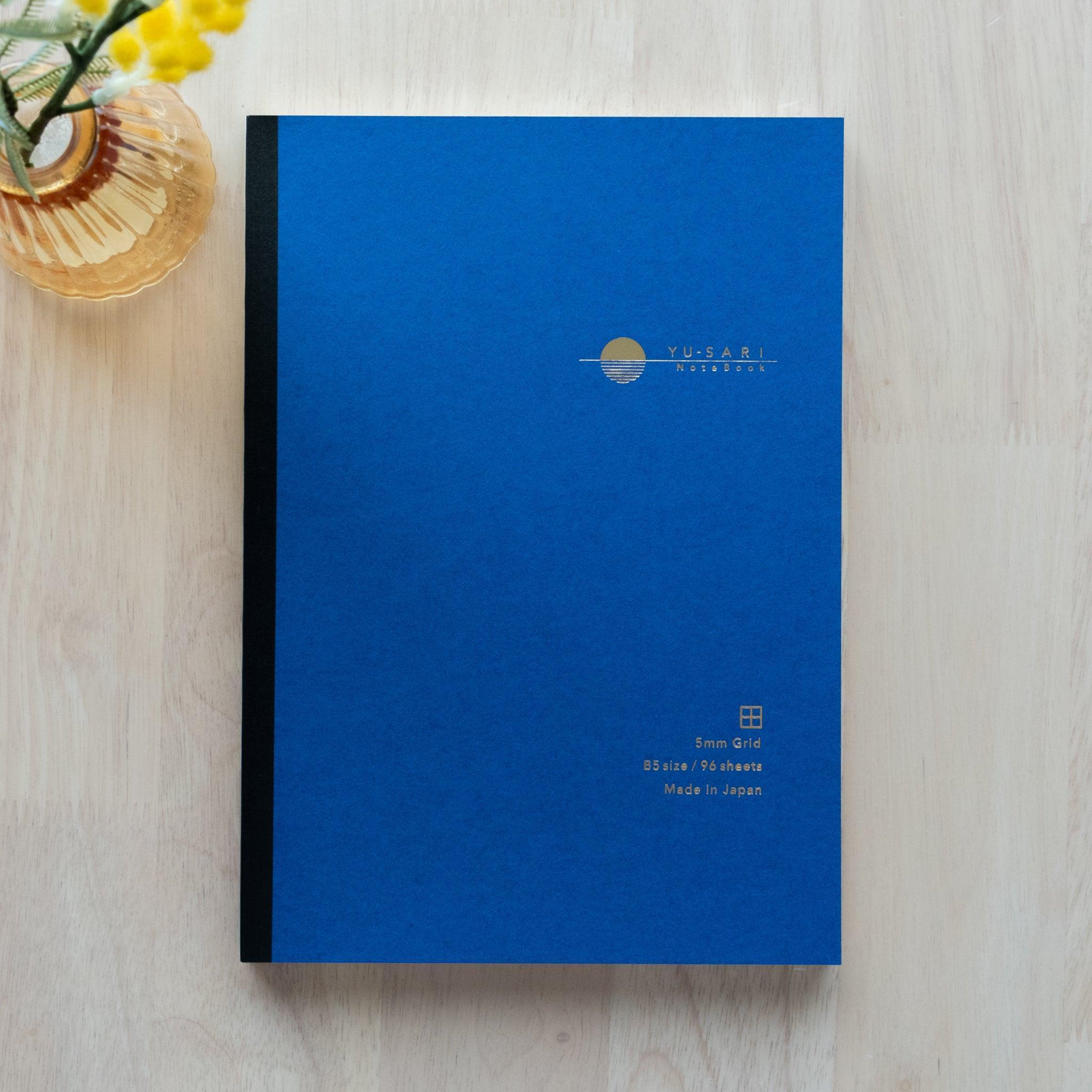 A blue YU-SARI notebook with gold embossing on the cover, featuring 5mm grid pages and A5 size, beautifully displayed next to a vase with yellow flowers.