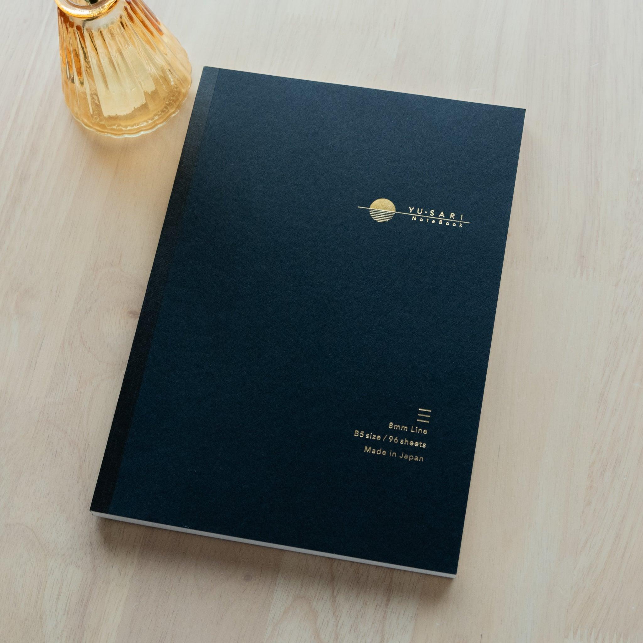 black notebook cover with gold-embossed "YU-SARI Notebook" logo, a minimal sun design, and specifications on a light wooden table.
