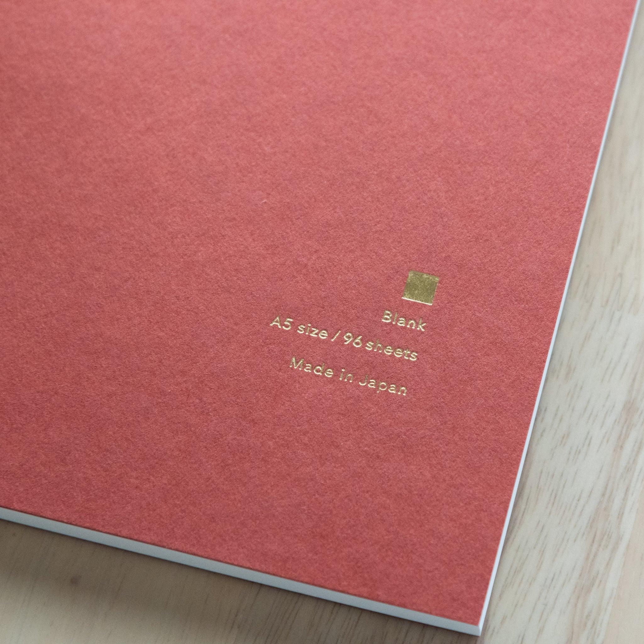 A blue YU-SARI notebook with gold embossing on the cover, featuring 5mm grid pages and A5 size, beautifully displayed next to a vase with yellow flowers.