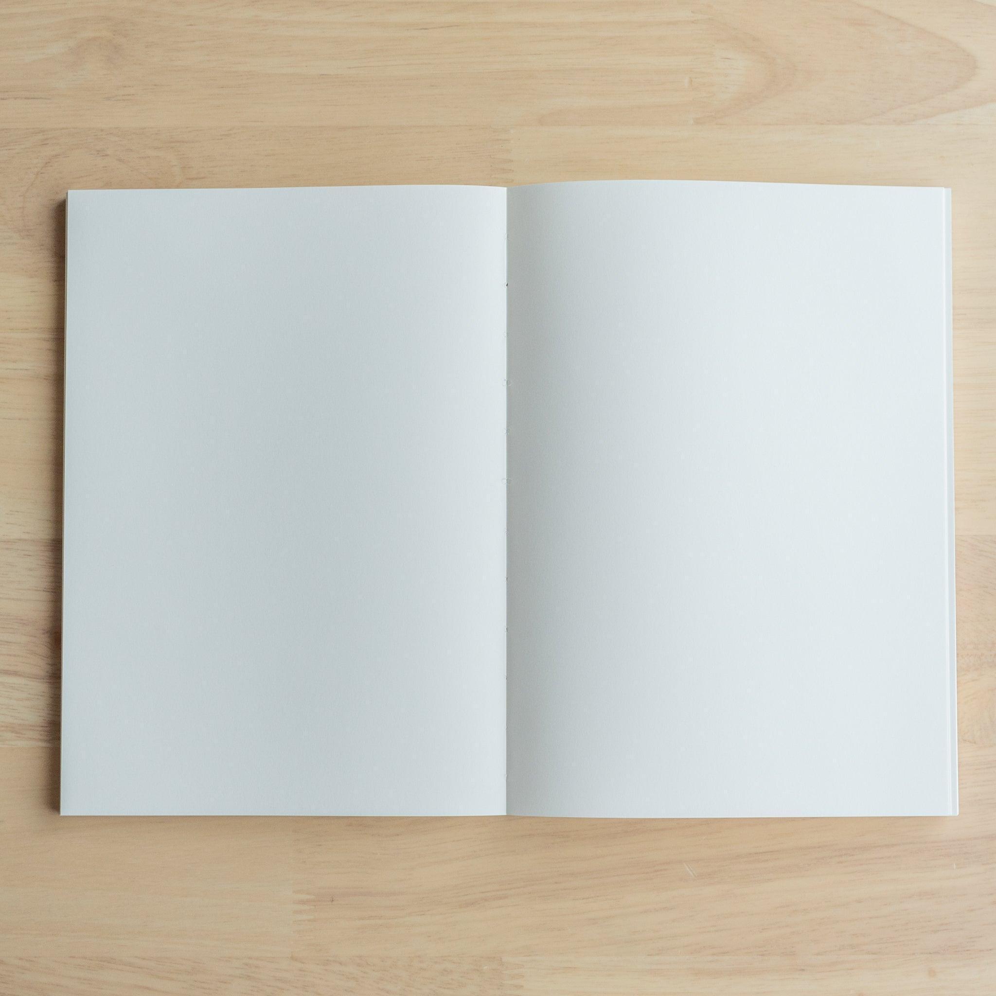 An open YU-SARI notebook revealing its smooth blank pages, ideal for writing or drawing, displayed on a wooden surface under soft light.