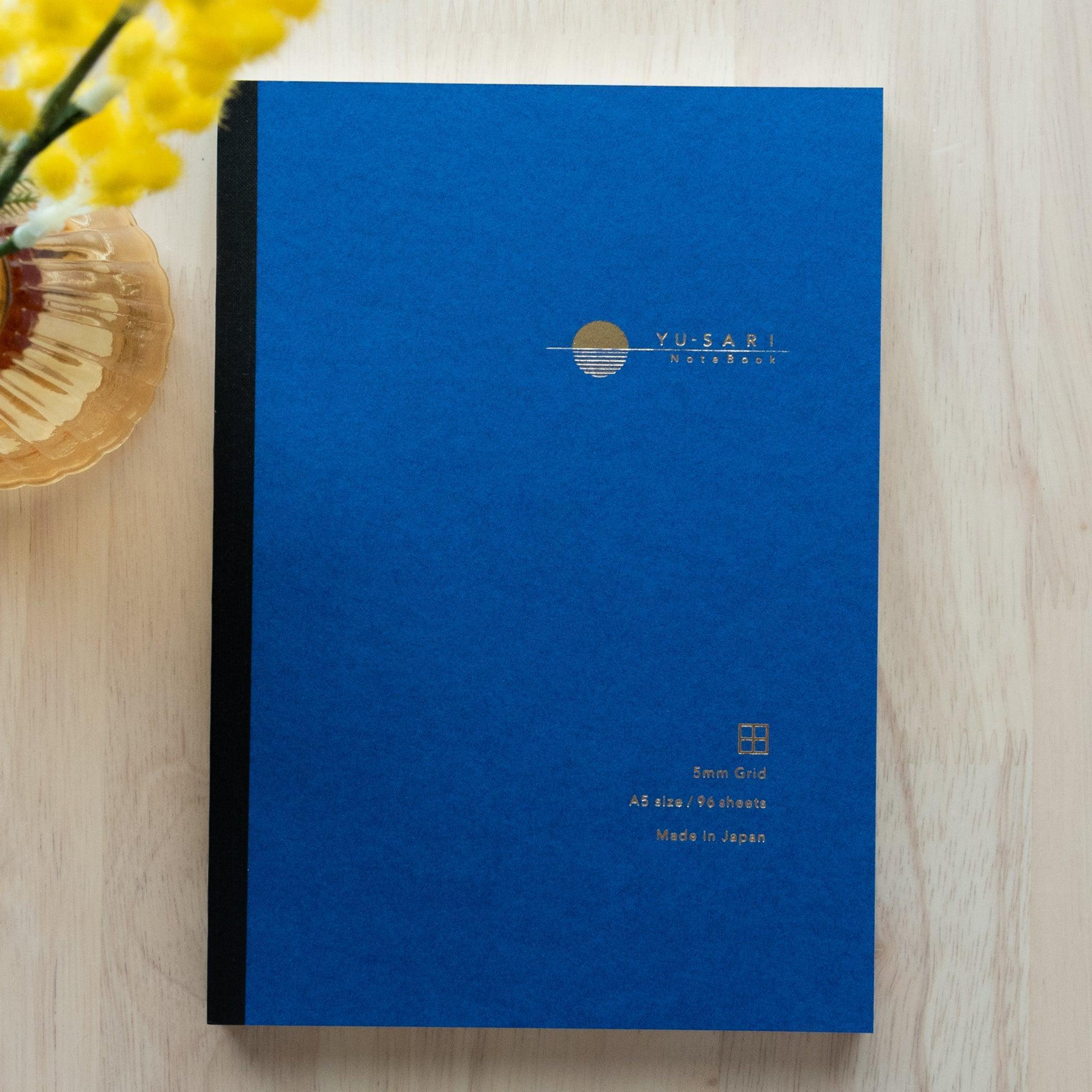 A blue YU-SARI notebook with gold embossing on the cover, featuring 5mm grid pages and A5 size, beautifully displayed next to a vase with yellow flowers.