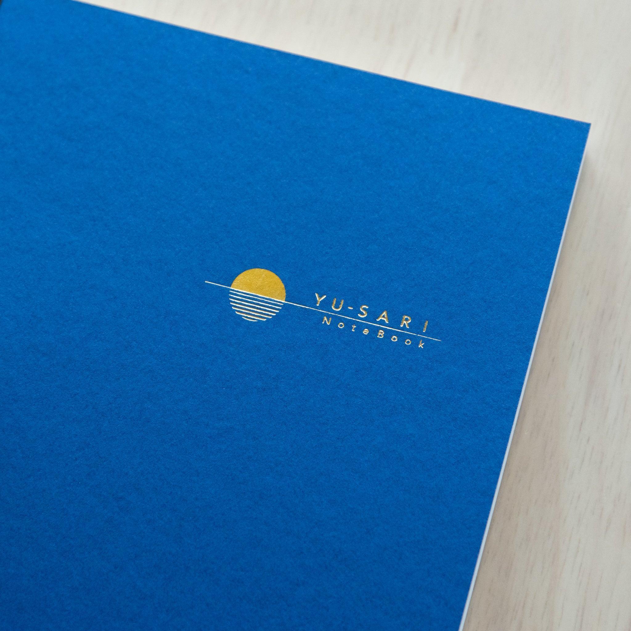 Three "Yu-Sari Notebooks" in red, blue, and black, displayed on a wooden table with a glass vase of yellow flowers.